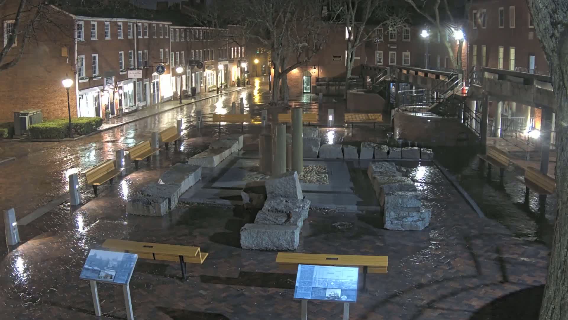 A wet, brick plaza at night features a stone structure with remnants of columns, surrounded by benches and illuminated by streetlights.