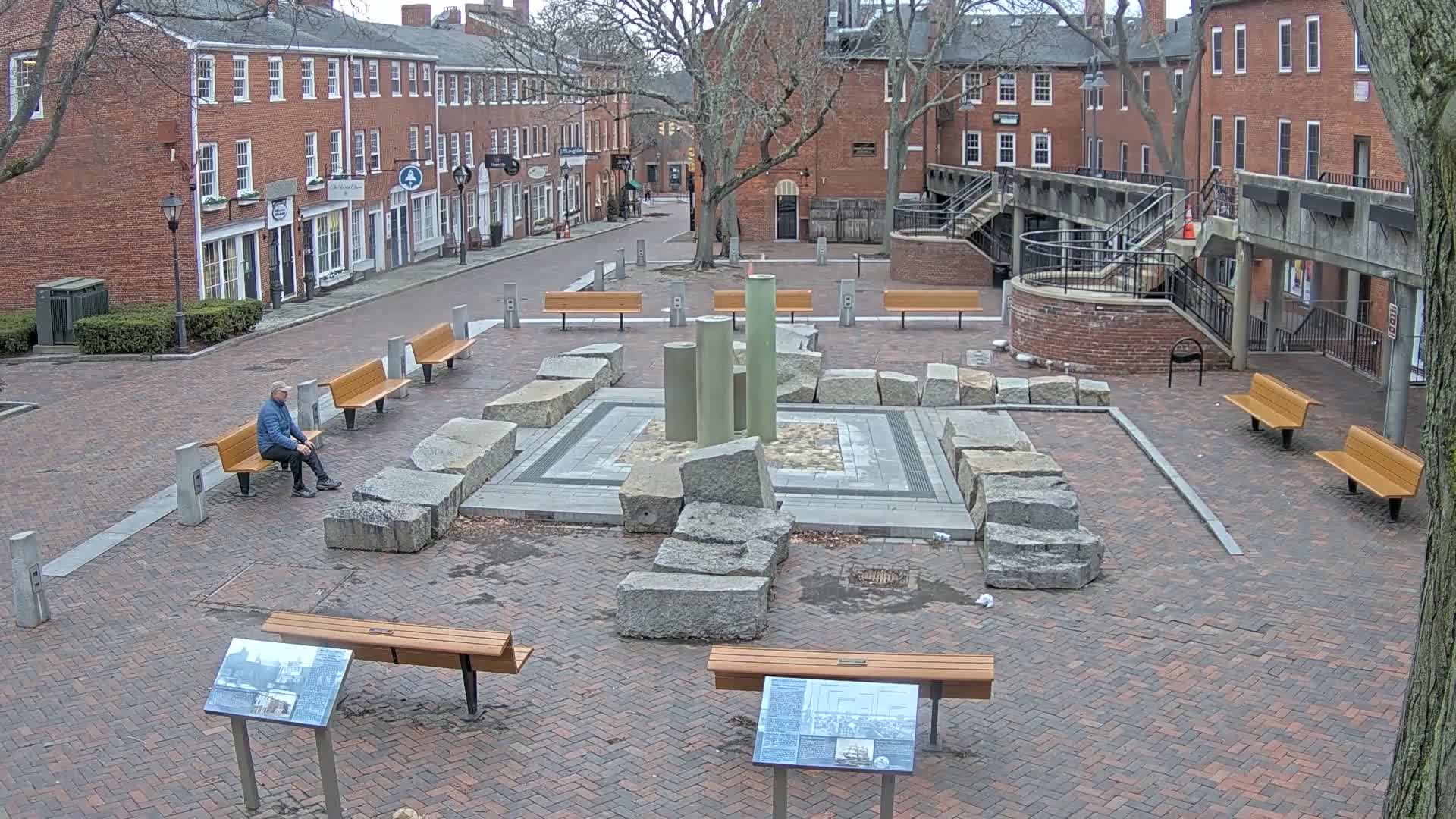 A man sits on a bench in a brick town square featuring a stone and column fountain, surrounded by more benches and brick buildings under a cloudy sky.