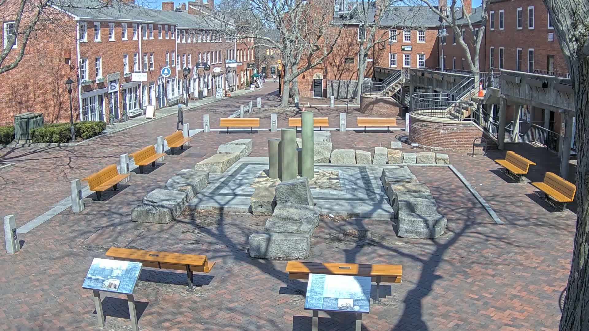 A brick plaza with several benches and a stone and metal fountain-like structure is surrounded by brick buildings under a sunny sky.