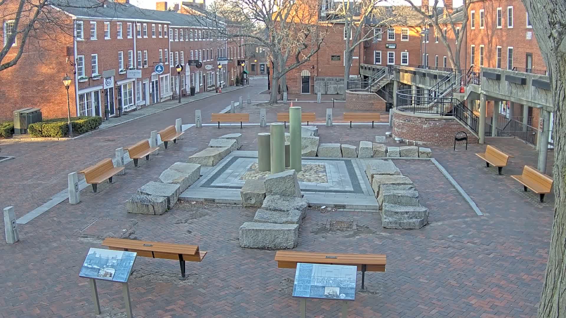 A brick plaza with several benches surrounds a stone fountain and informational plaques under a mostly clear sky.