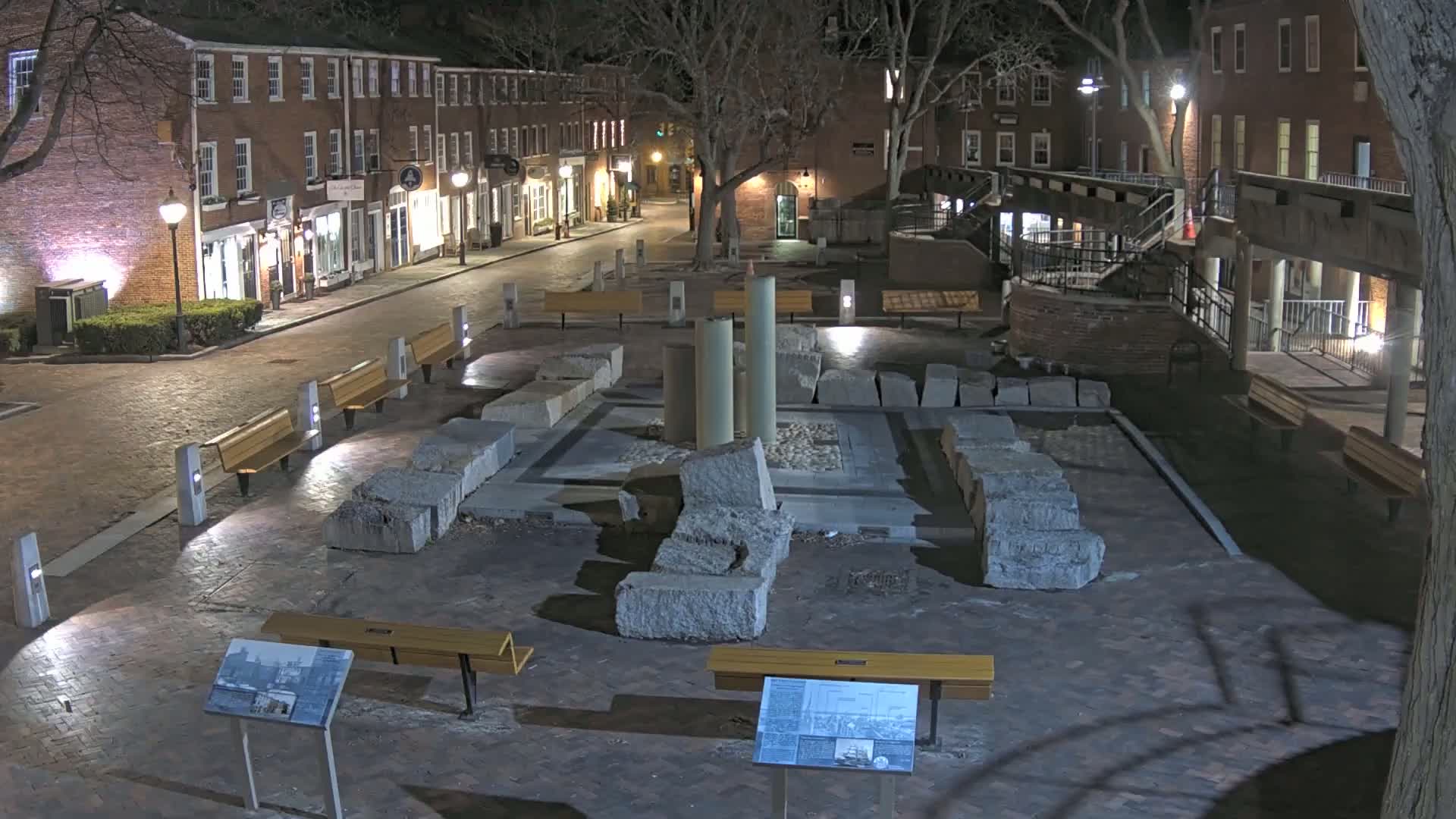 A nighttime view of a brick-paved town square with several benches, a central stone structure resembling ruins, and surrounding buildings under the cover of trees.