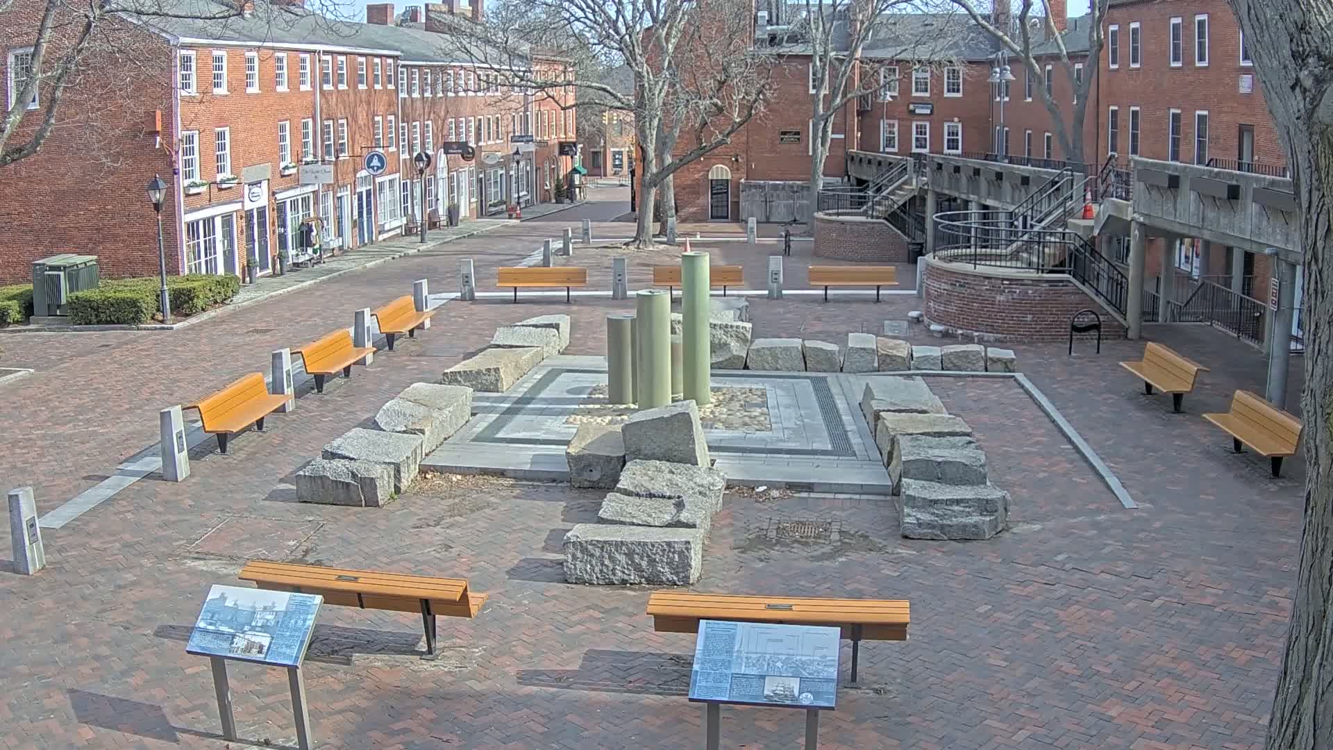 A brick plaza with several benches surrounds a stone fountain, under a sunny sky.