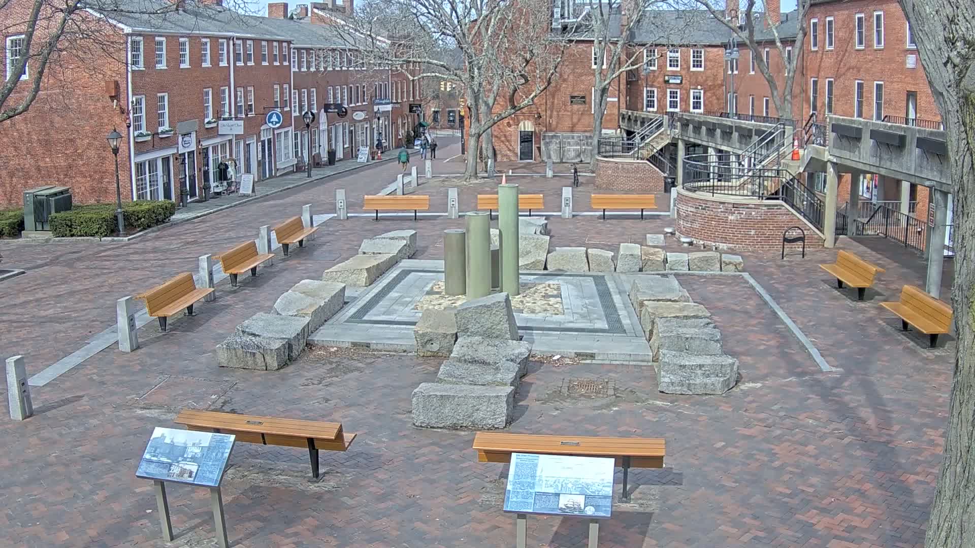 A brick plaza with benches, a stone fountain, and several brick buildings in the background on a sunny day.