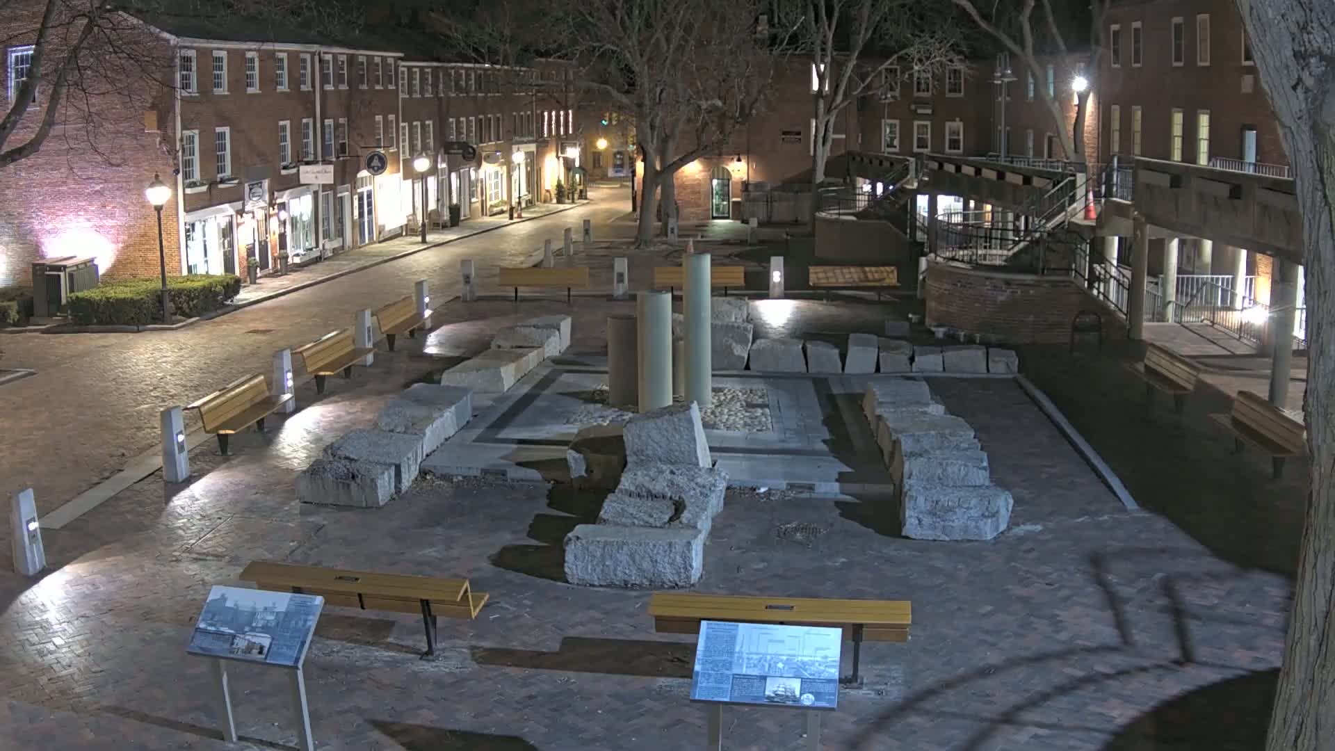 A nighttime view of a brick-paved plaza with a central stone structure, several benches, and surrounding brick buildings under a dark sky.