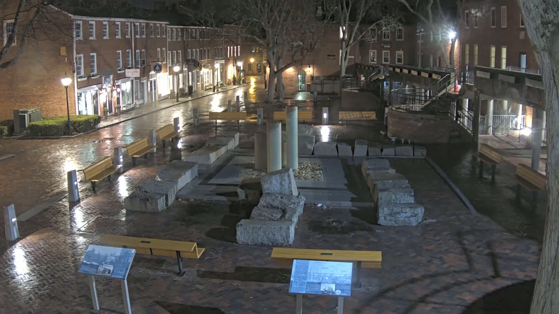 A rain-slicked brick plaza at night features a stone monument, several benches, and is flanked by brick buildings.
