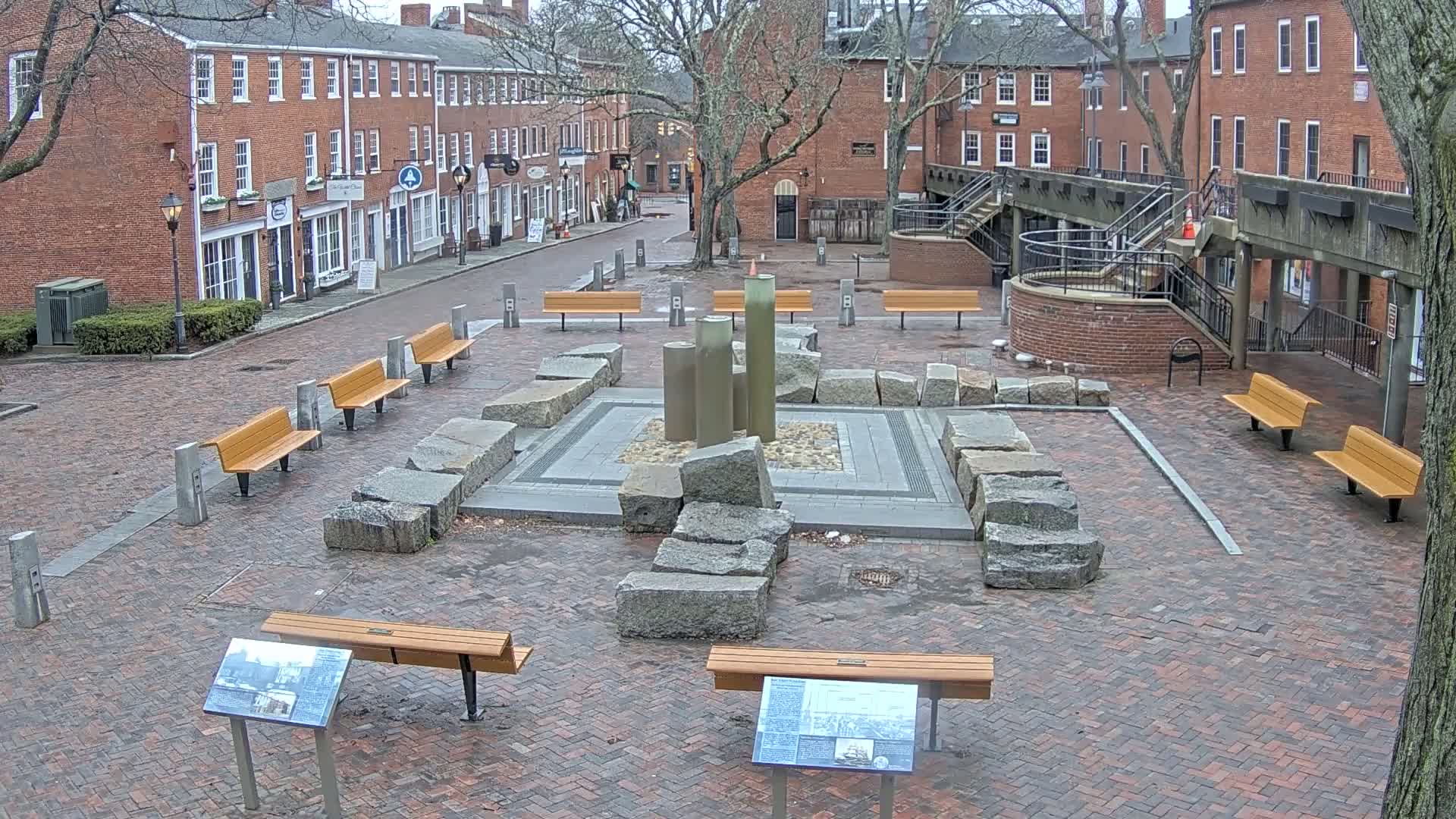 A brick plaza with several benches surrounds a stone fountain, and brick buildings line the street behind it on an overcast day.