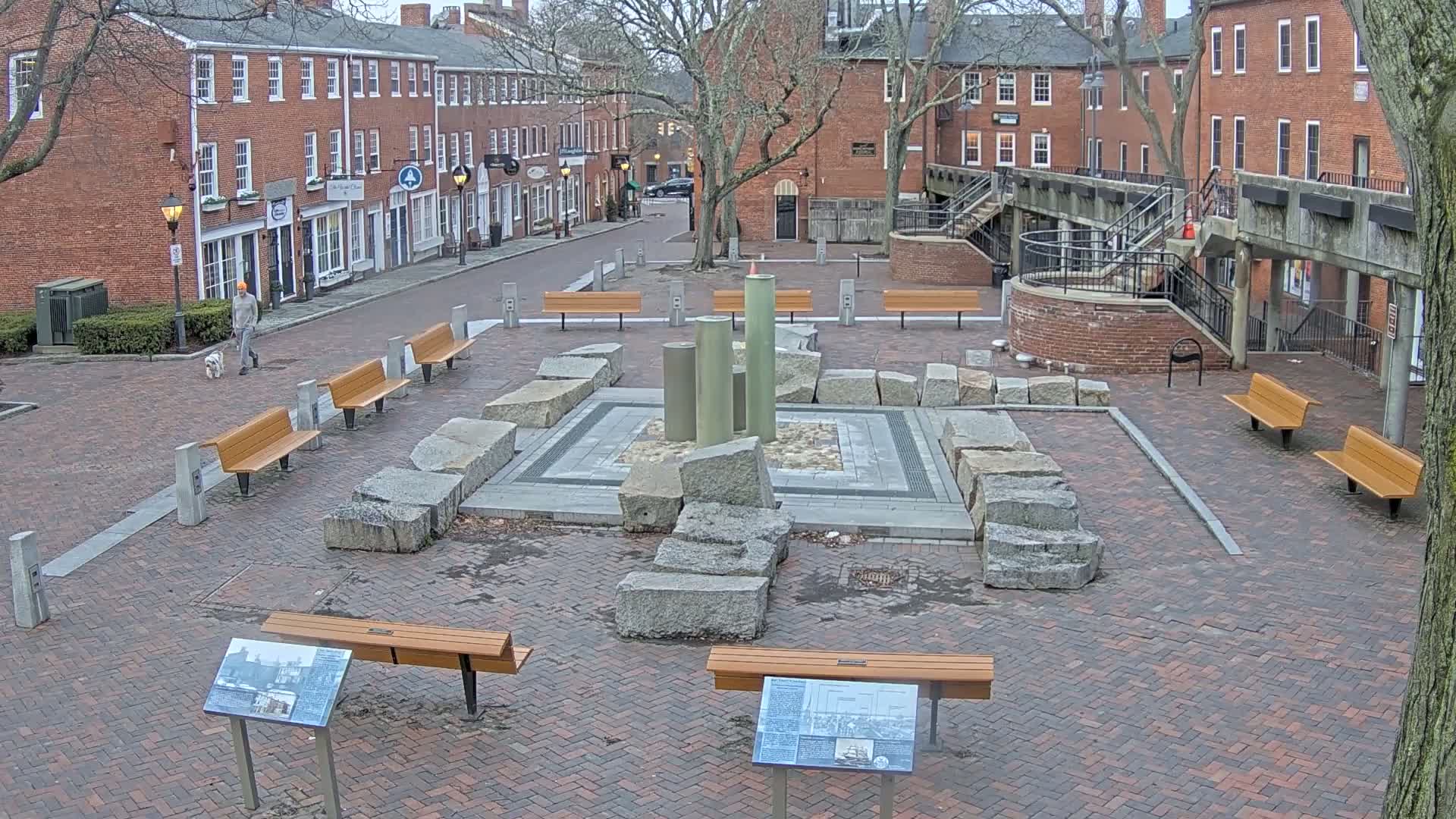 A brick plaza with several benches surrounds a stone and metal fountain, and a person walks a dog down a street lined with brick buildings in an overcast setting.