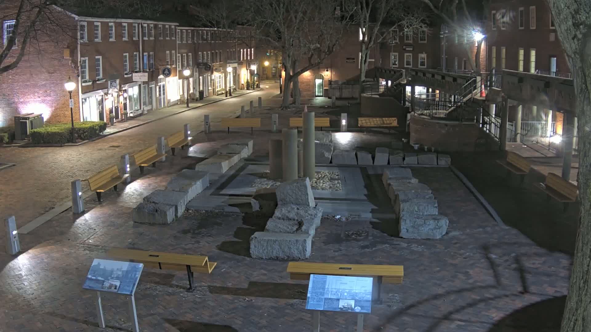 At night, a brick-paved plaza features a stone monument with several columns, surrounded by benches and illuminated by streetlights, with brick buildings lining the street in the background.