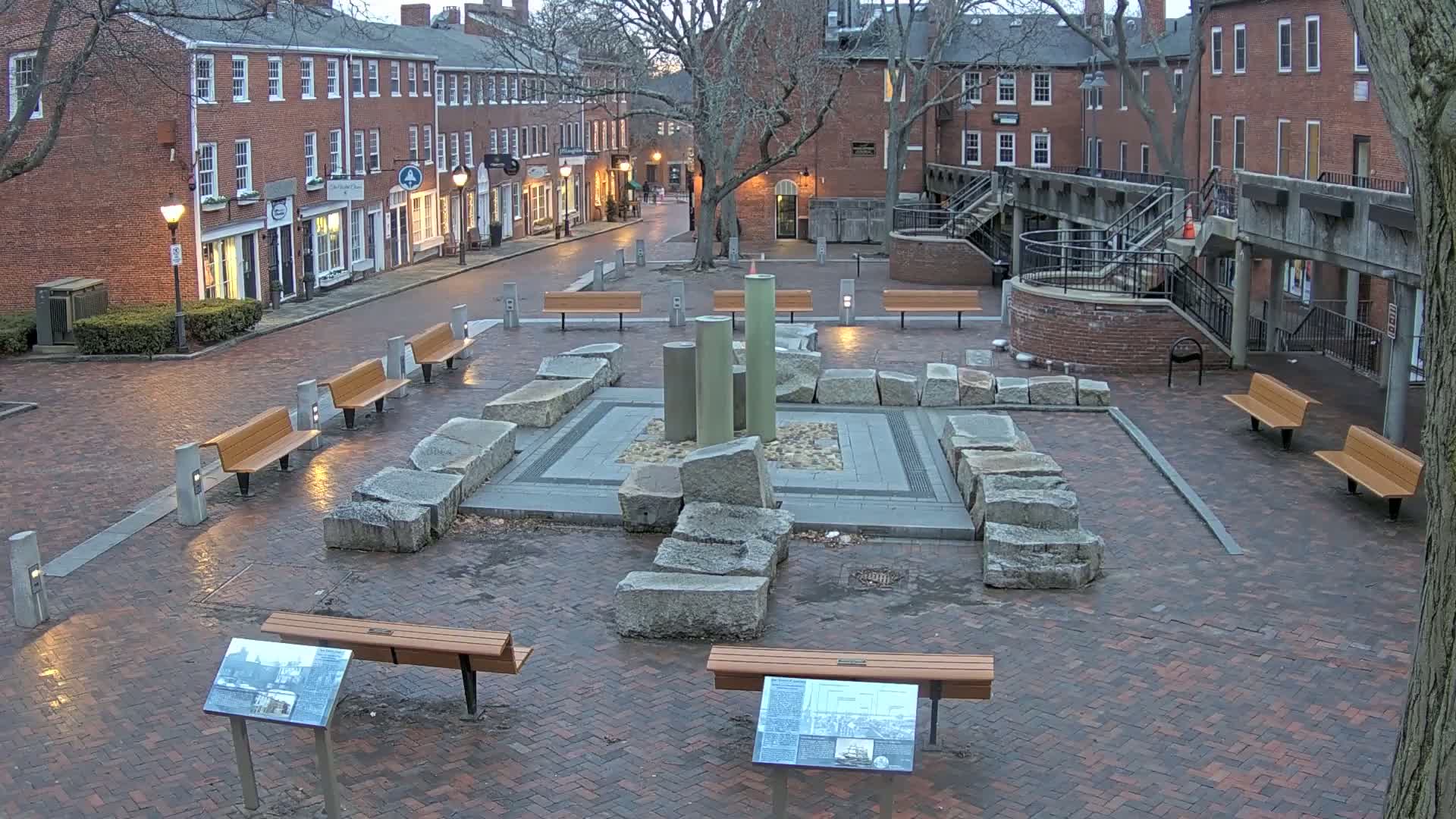 A brick plaza, illuminated at dusk, features a central stone structure with tall, cylindrical elements, surrounded by several benches and informational plaques.