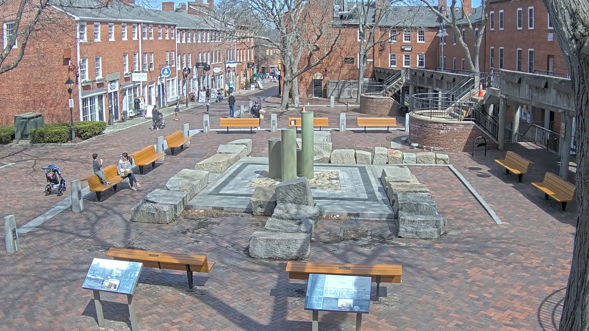 A brick plaza with benches, a stone and metal fountain, and several people sitting on benches on a sunny day.