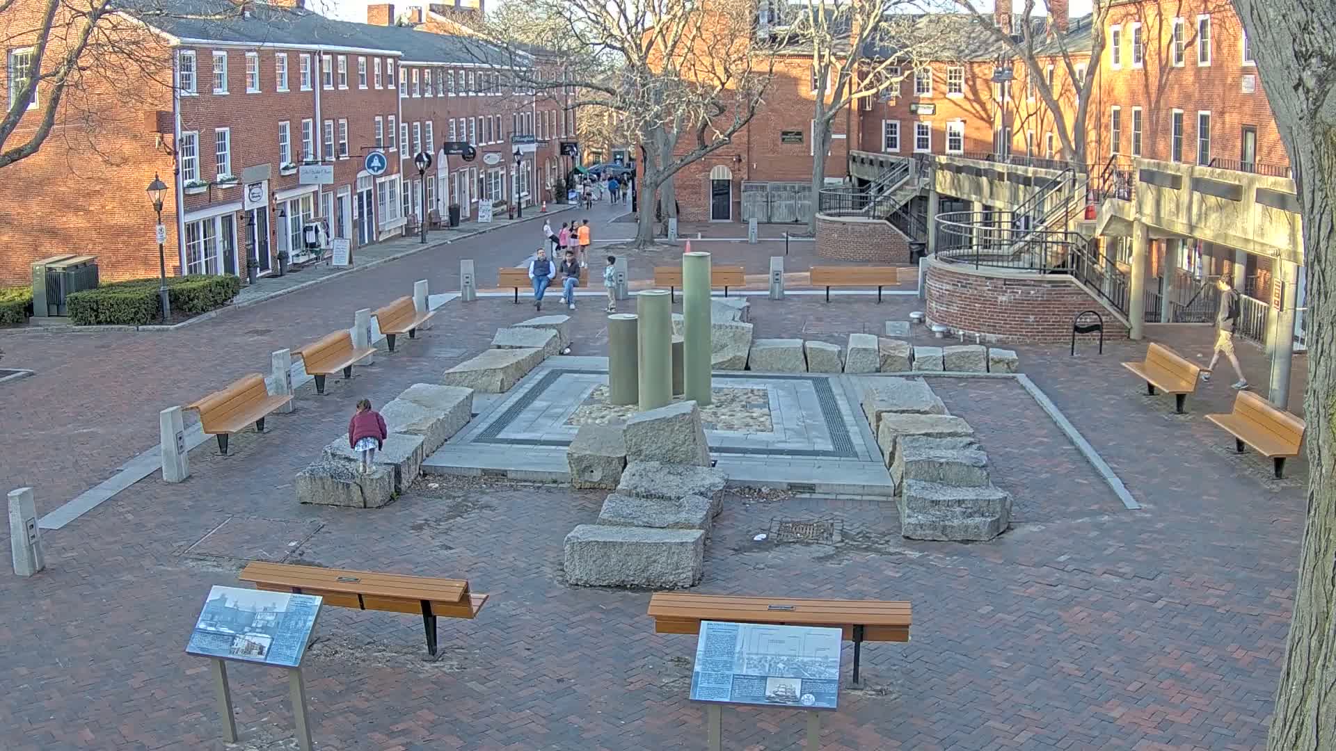 A brick town square with several benches, a stone fountain, and brick buildings surrounding it on a sunny day.