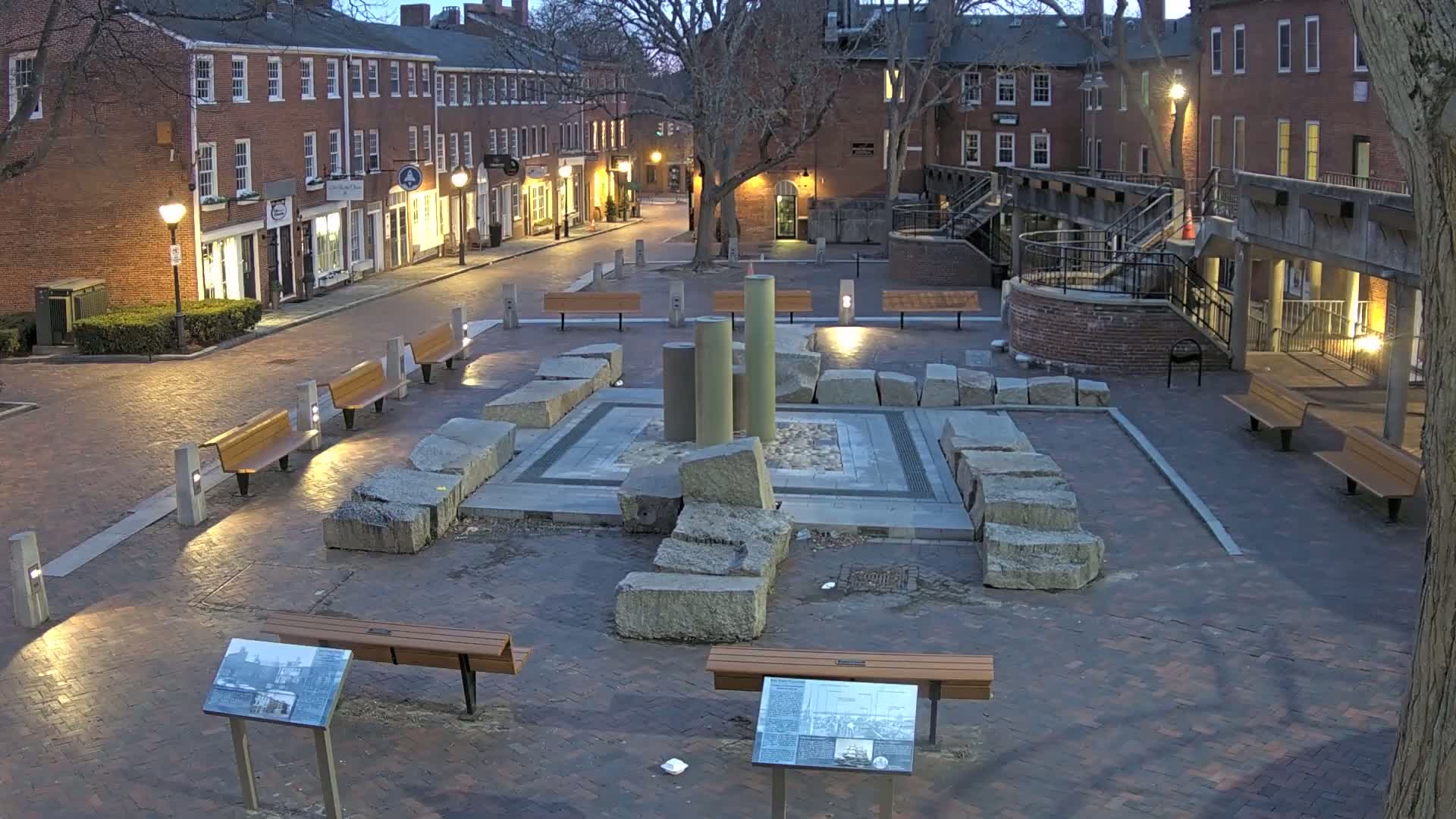 A brick plaza at twilight, featuring a central stone structure with several columns, surrounded by benches and lit by streetlights, bordered by brick buildings.