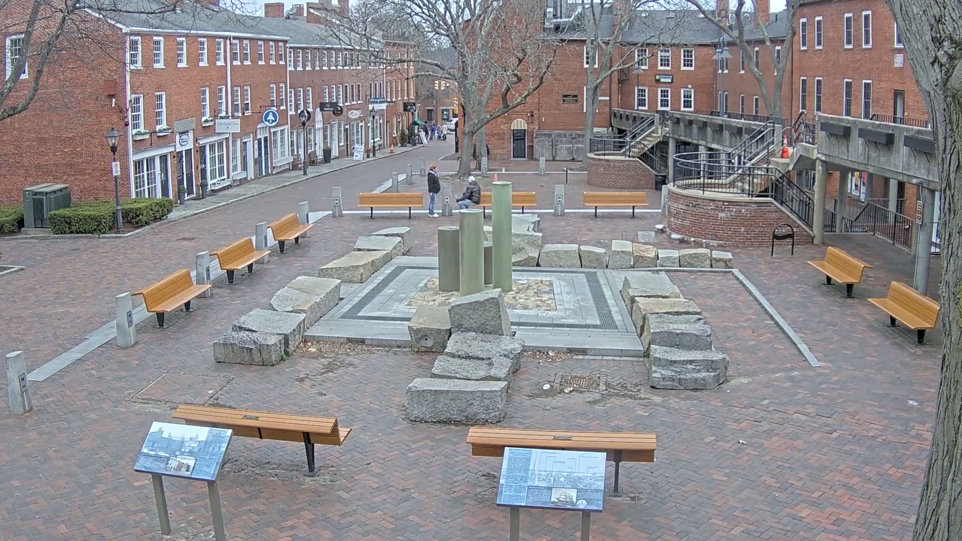 A brick plaza with several benches surrounding a stone structure and two people standing near a fountain, under a mostly cloudy sky.