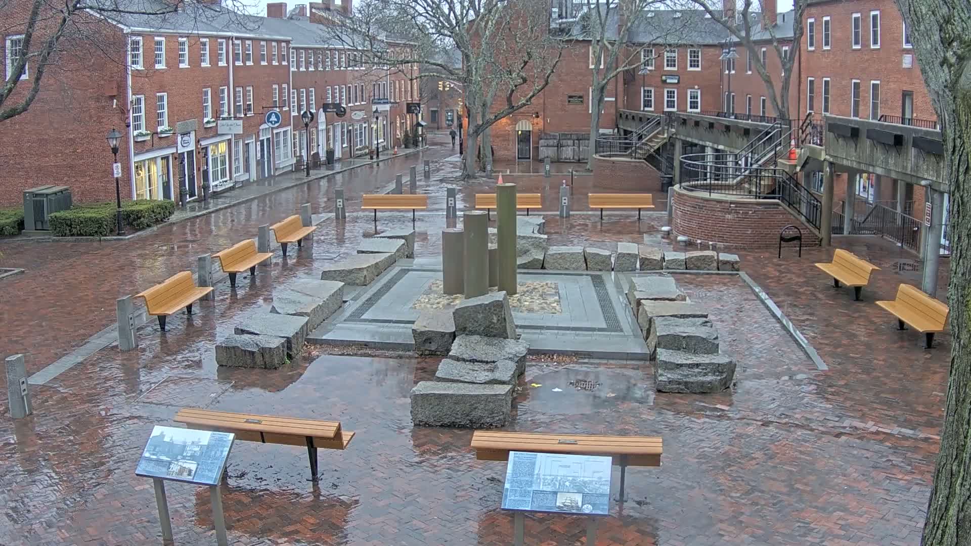 A wet brick plaza with several benches surrounding a stone fountain, situated between brick buildings on an overcast day.