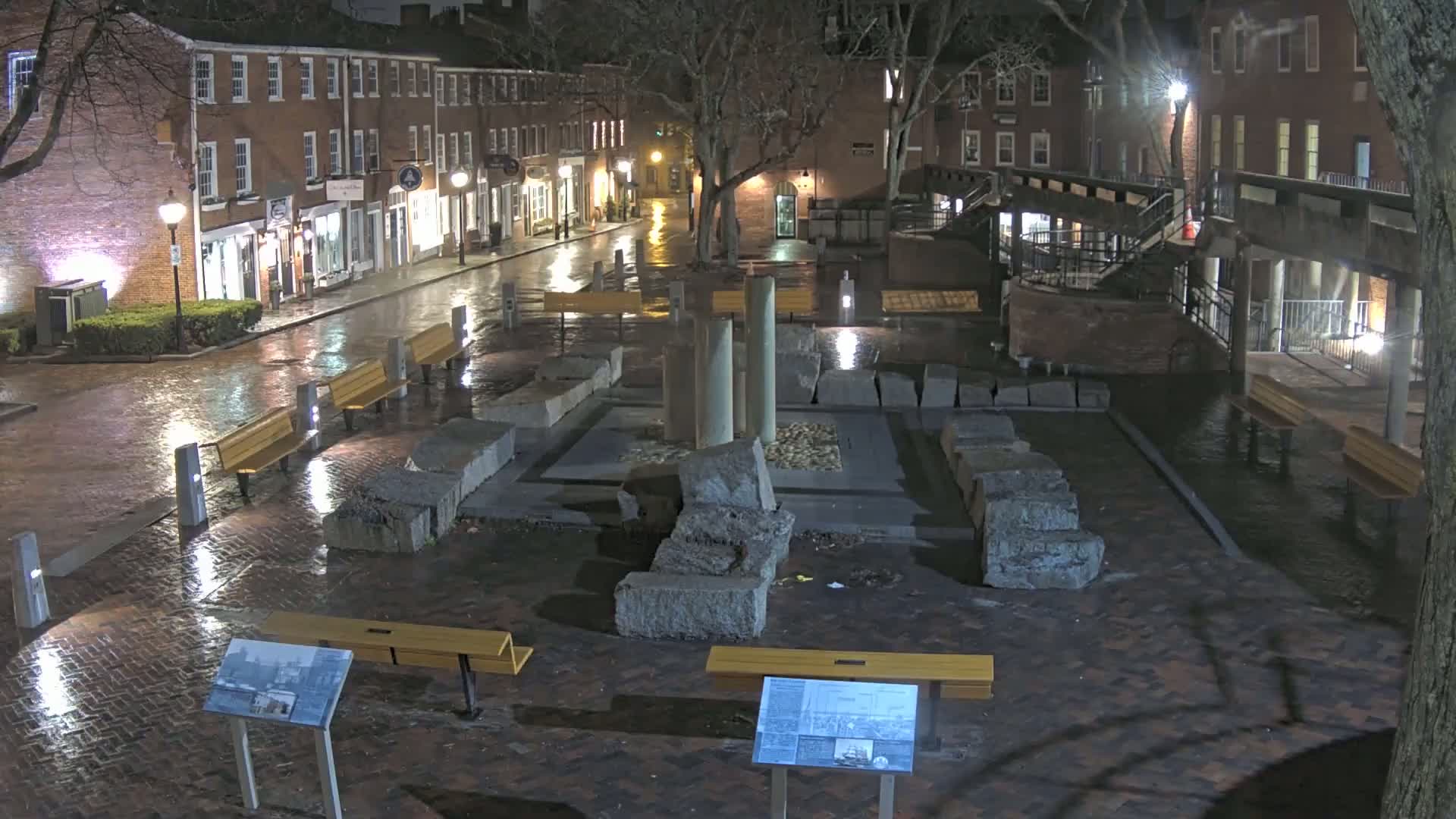 A rain-slicked brick plaza at night features a stone monument, several benches, and is bordered by lit buildings.