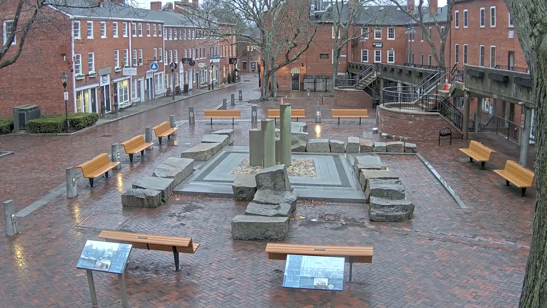 A brick plaza with several benches and a stone fountain is wet from recent rain, surrounded by brick buildings.