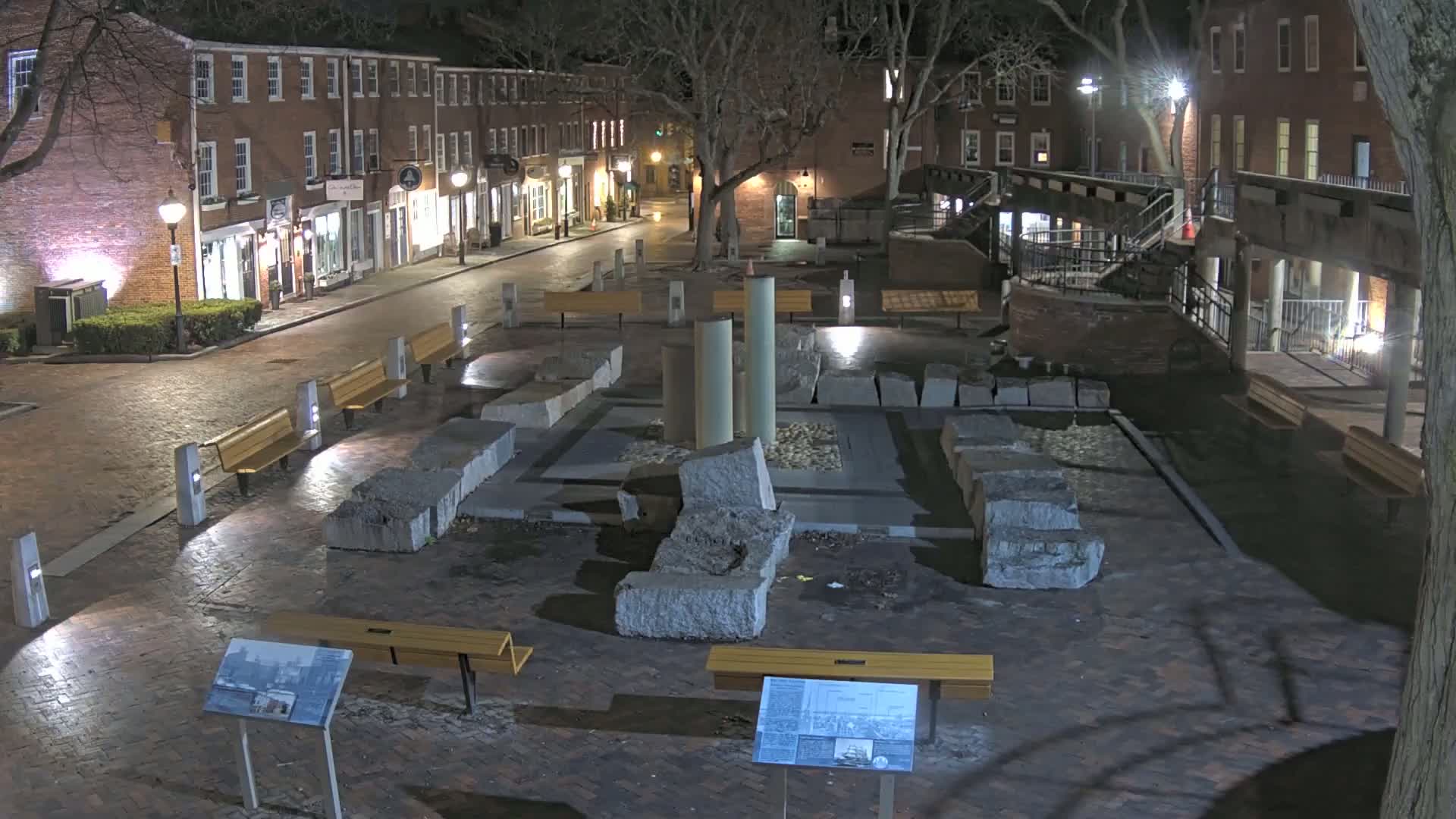 At night, a brick-paved town square contains a stone monument and several benches, surrounded by brick buildings.