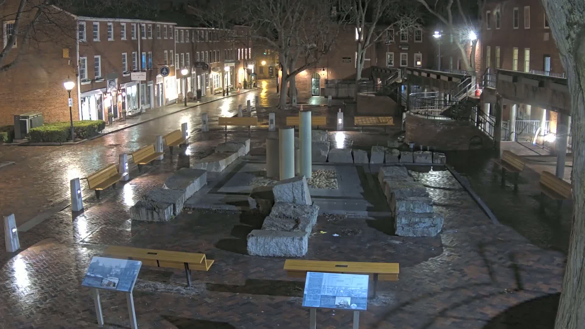 A wet brick plaza at night, featuring several benches and a central stone structure, is surrounded by brick buildings with illuminated windows.