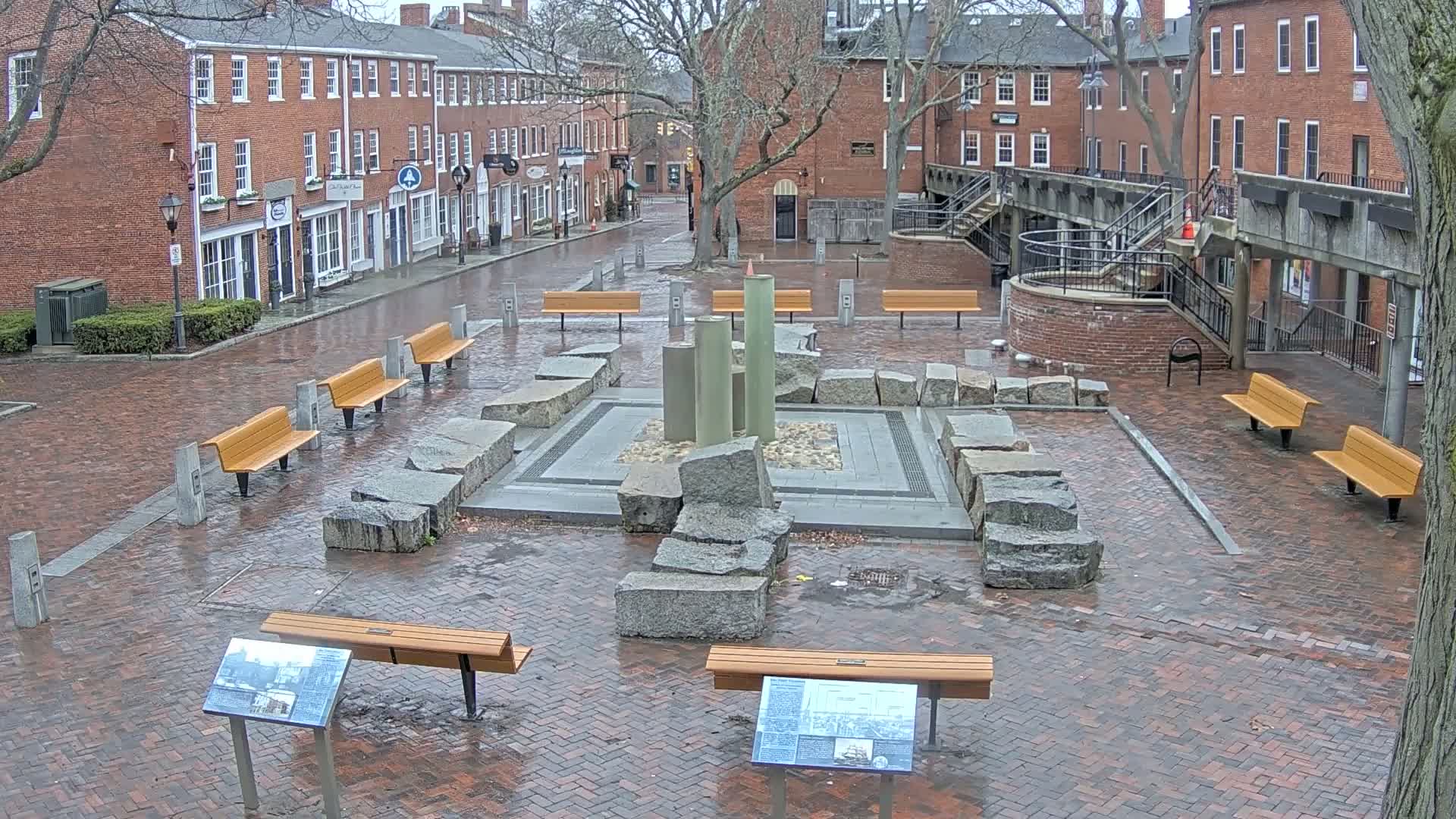A brick plaza with several benches, a stone fountain, and informational plaques, surrounded by brick buildings under a rainy sky.
