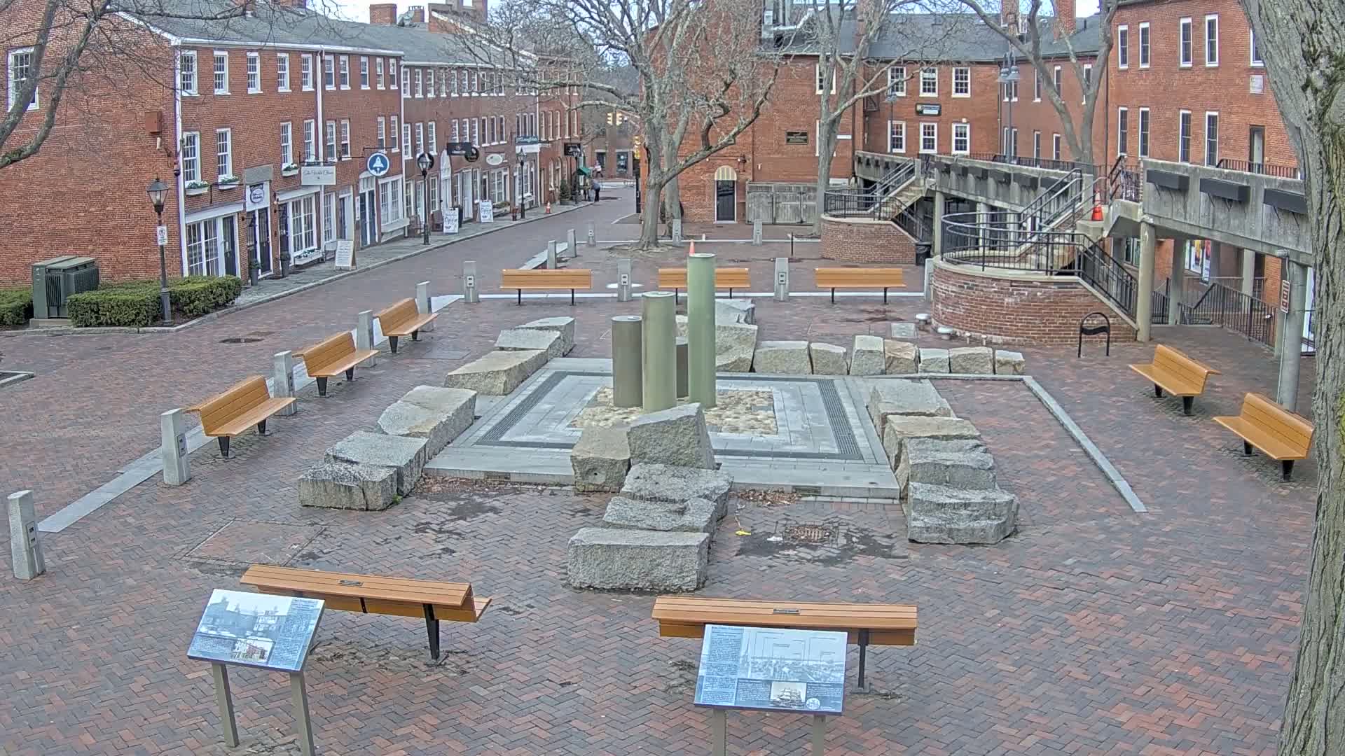 A brick plaza with several benches surrounds a stone and metal fountain, situated between brick buildings under a clear, daytime sky.