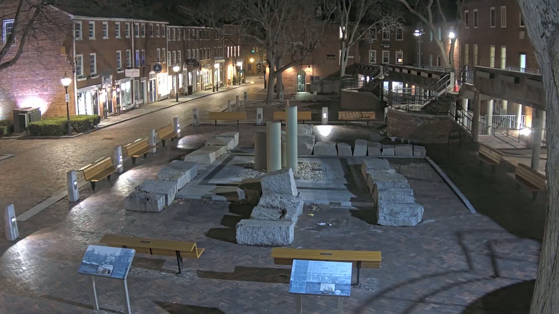 A nighttime view of a brick plaza featuring large stone remnants and several benches, surrounded by illuminated brick buildings.