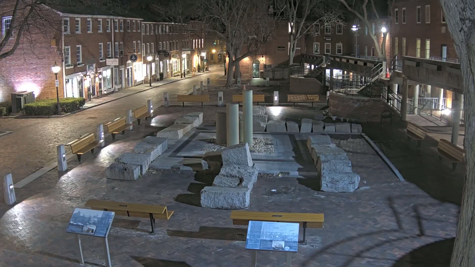 At night, a brick-lined town square features a central display of large stone blocks and columns, surrounded by several benches under ambient lighting.