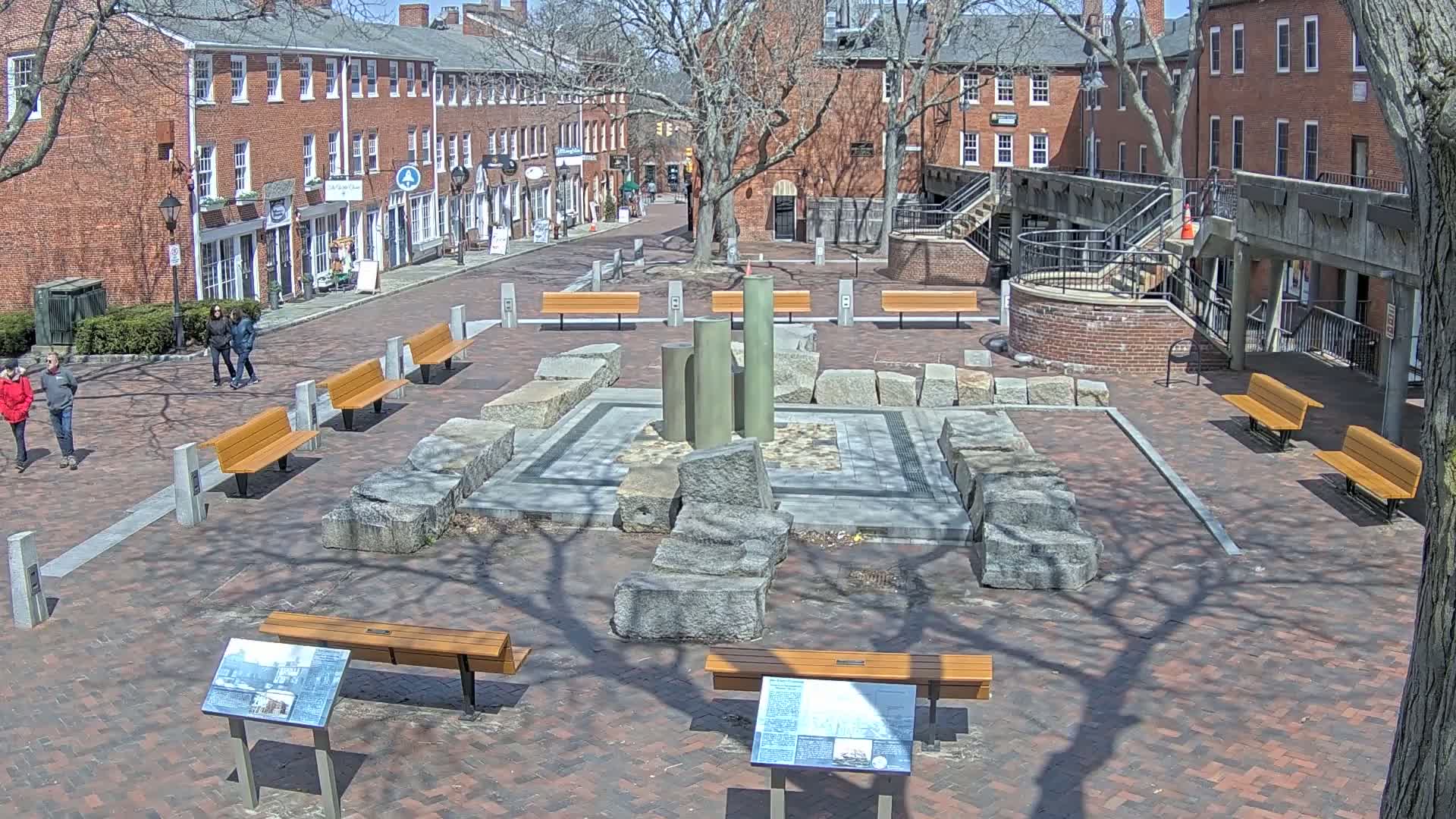 A brick plaza with several benches, a stone structure, and surrounding brick buildings on a sunny day.