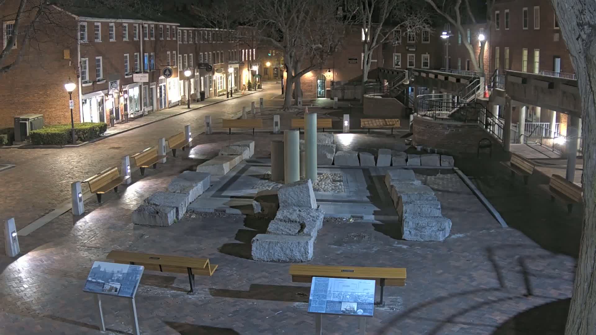 A nighttime view of a brick-paved plaza with stone ruins at its center, surrounded by benches and brick buildings under a clear, dark sky.