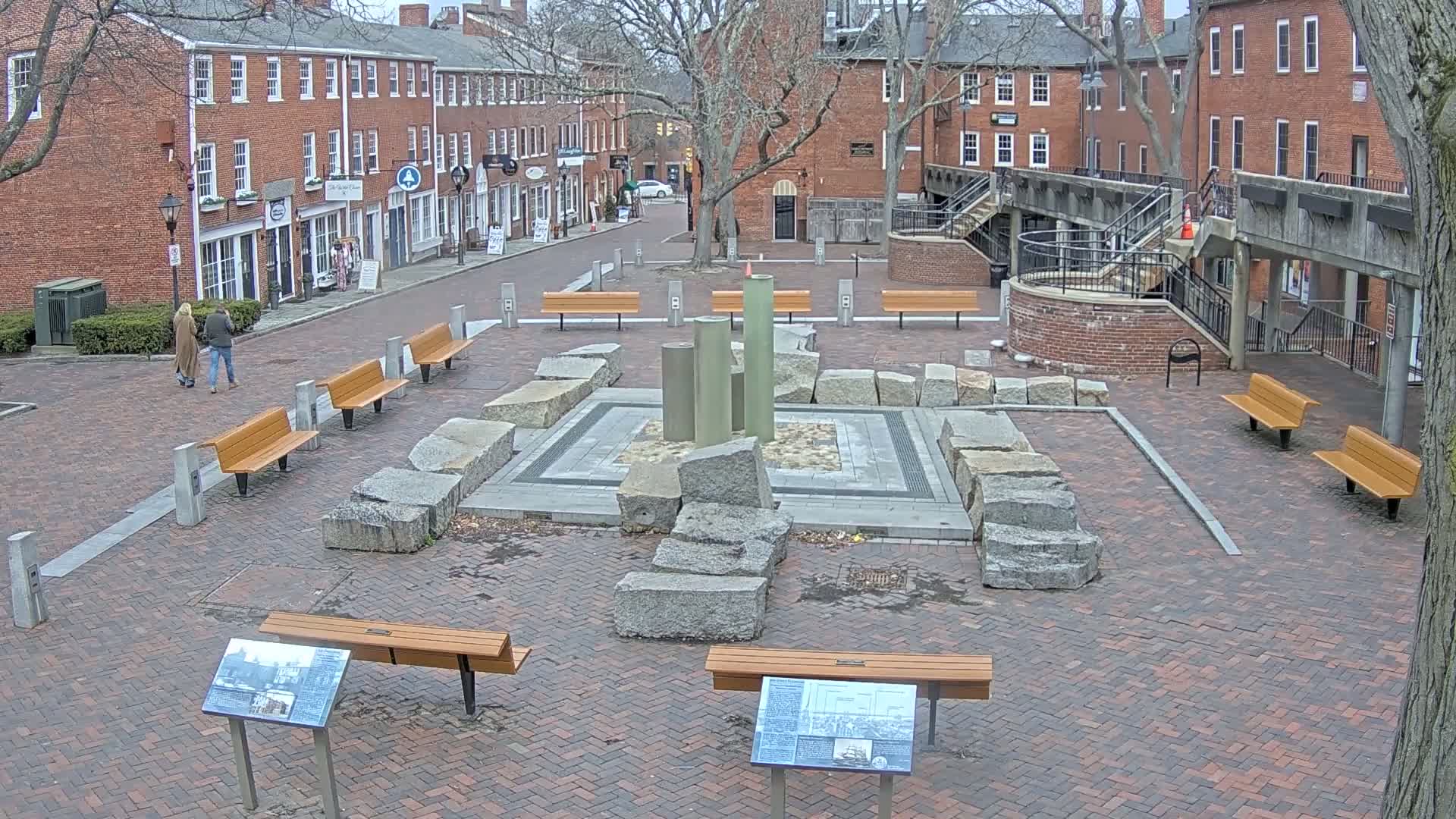 A brick plaza with several benches surrounds a central fountain made of large, rough-hewn stones and several tall, cylindrical structures, all under a cloudy sky.
