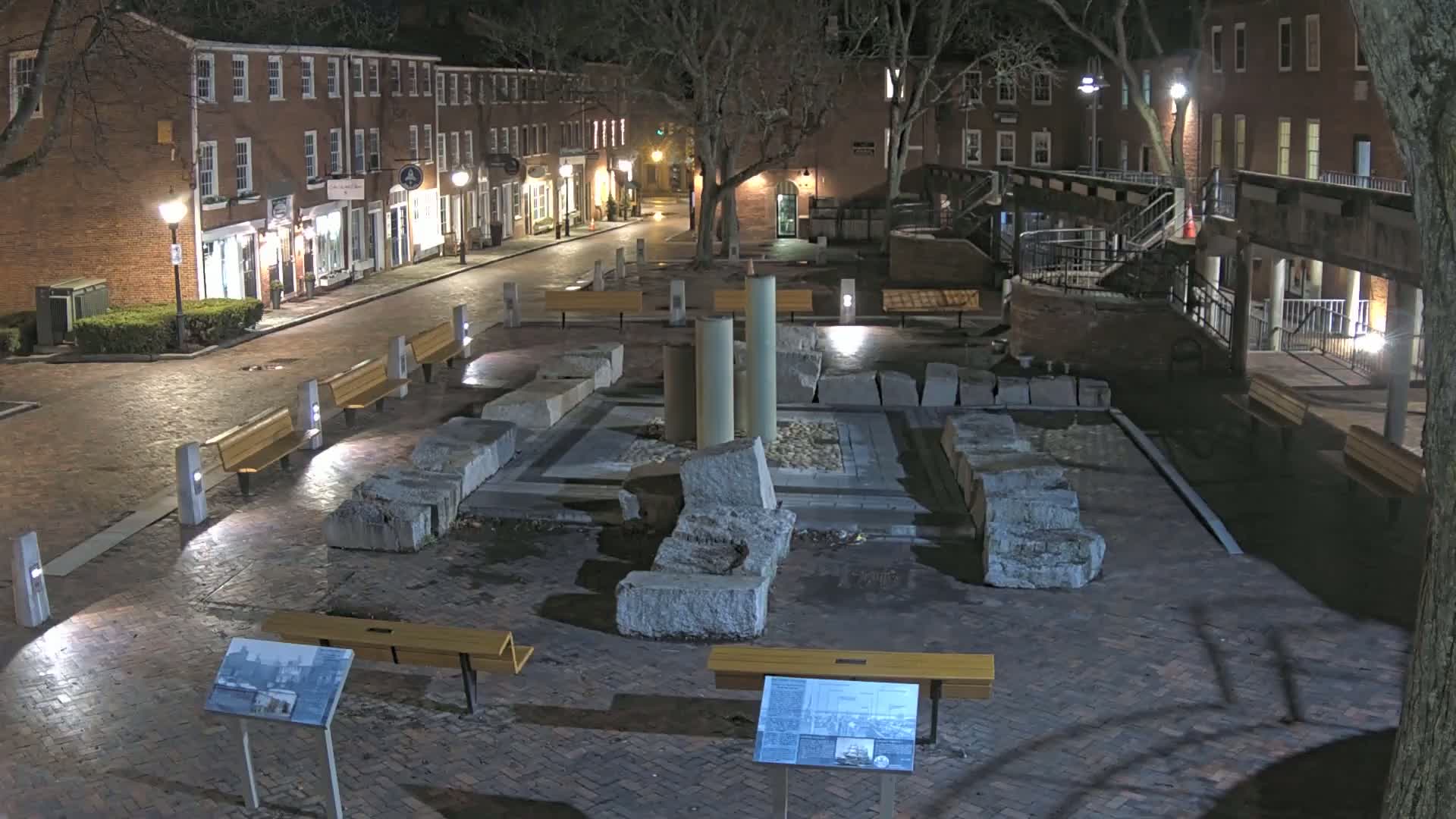 A nighttime view of a brick plaza with a stone ruin at its center, surrounded by benches and buildings with illuminated windows.