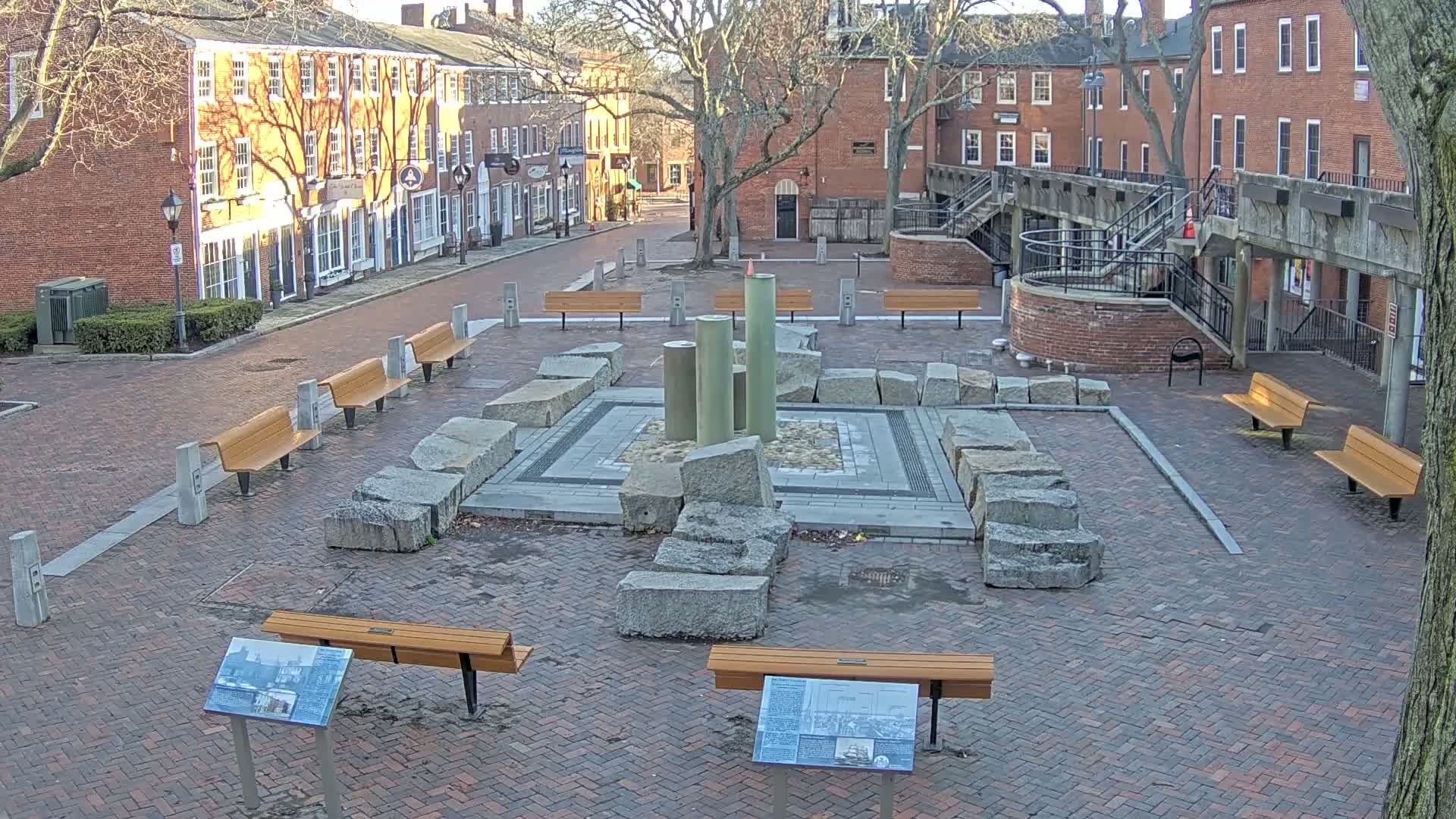 A brick plaza with several benches surrounds a stone fountain featuring tall, cylindrical structures, all under a clear, sunny sky.