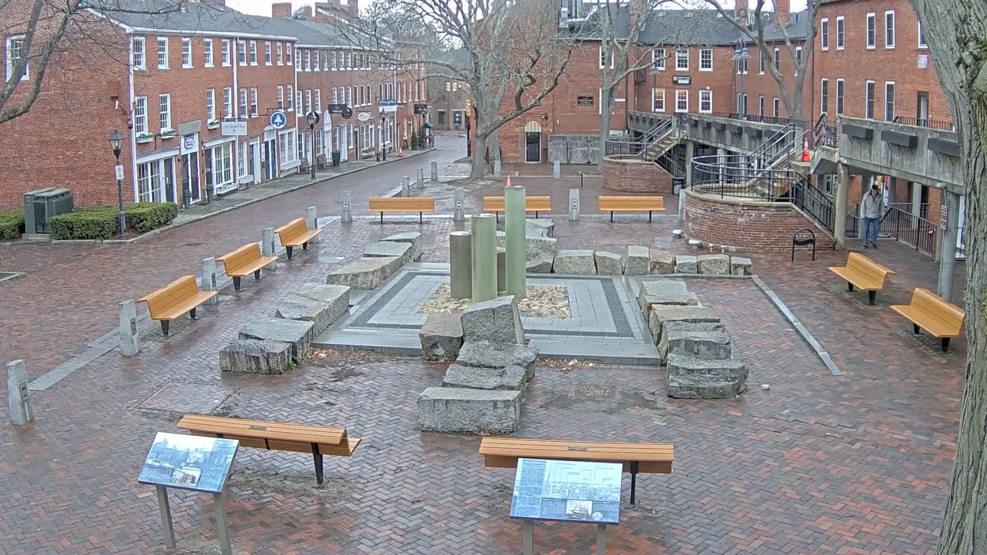 A brick plaza with several benches surrounds a stone and metal fountain, with brick buildings and leafless trees visible in the background under an overcast sky.