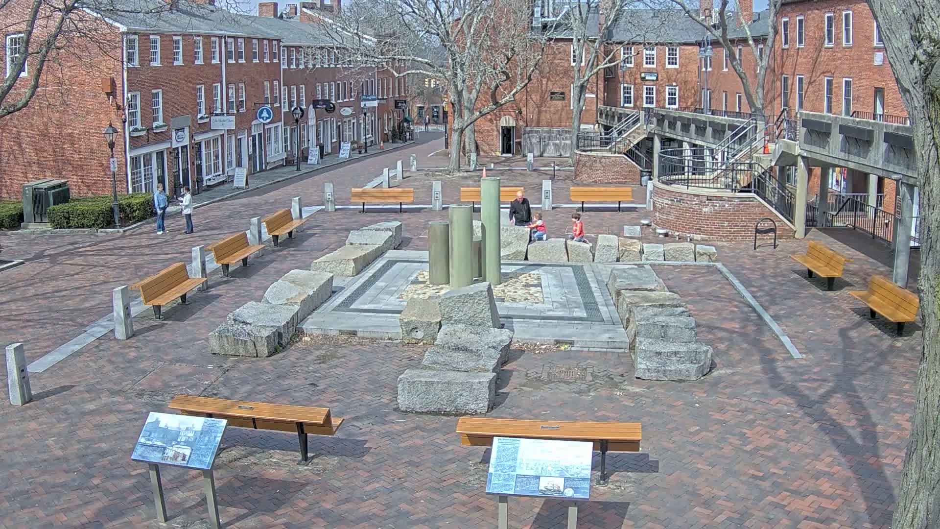 A brick plaza with benches, a stone fountain, and several people, surrounded by brick buildings under a sunny sky.