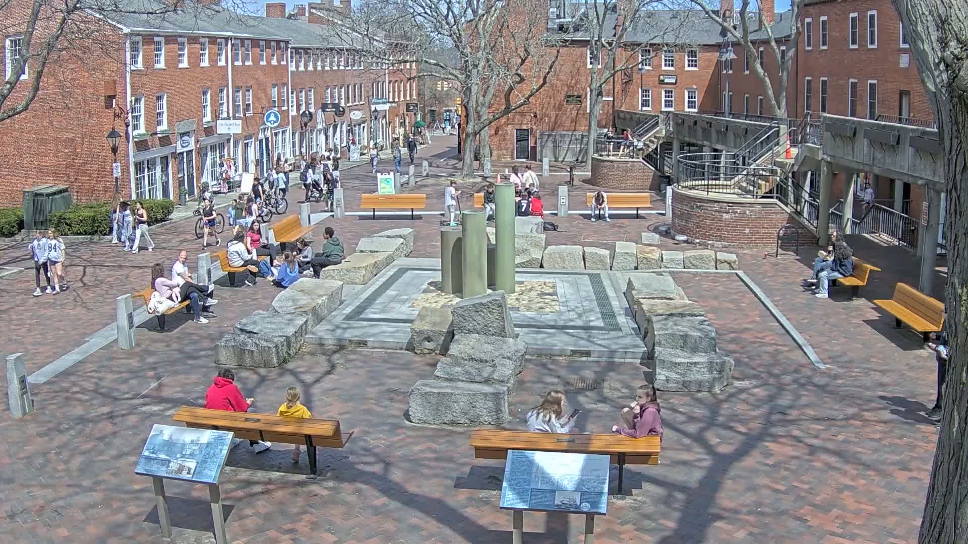 A brick plaza with a stone fountain, benches, and people sitting and walking under a sunny sky.