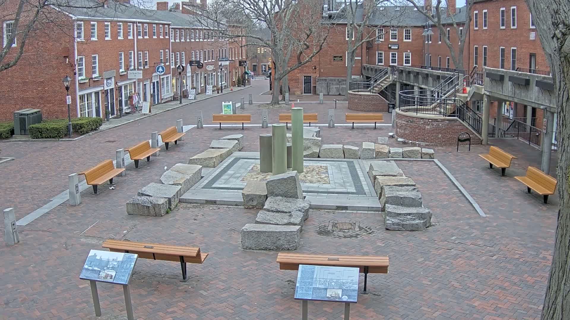 A brick plaza with several benches surrounds a central fountain constructed of large granite stones and metal pillars, under an overcast sky.