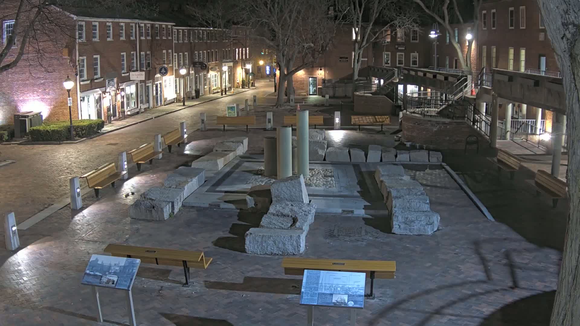 At night, a brick-paved plaza features large stone remnants in its center, surrounded by benches and streetlights, with a quiet street lined with brick buildings in the background.