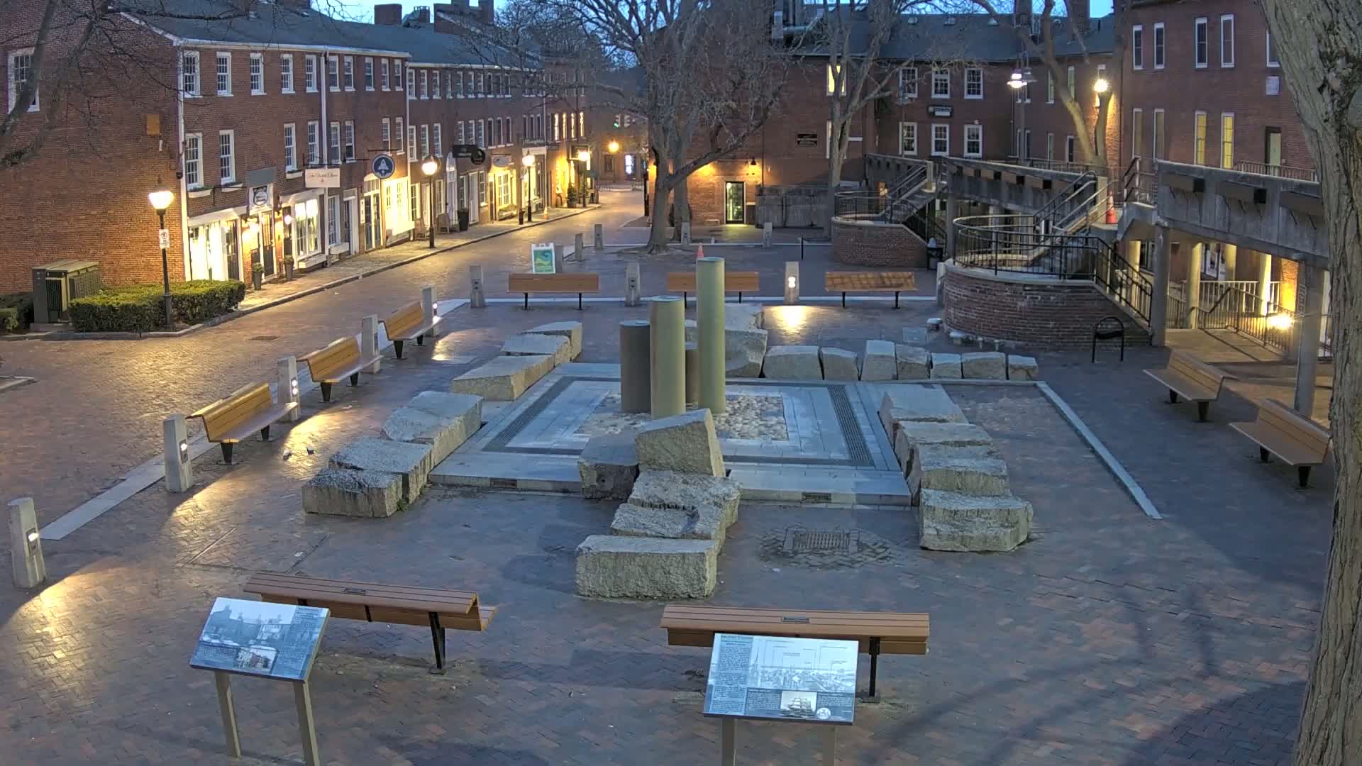 A brick-paved town square at twilight, featuring a central stone structure with columns, several benches, and buildings lining the square.