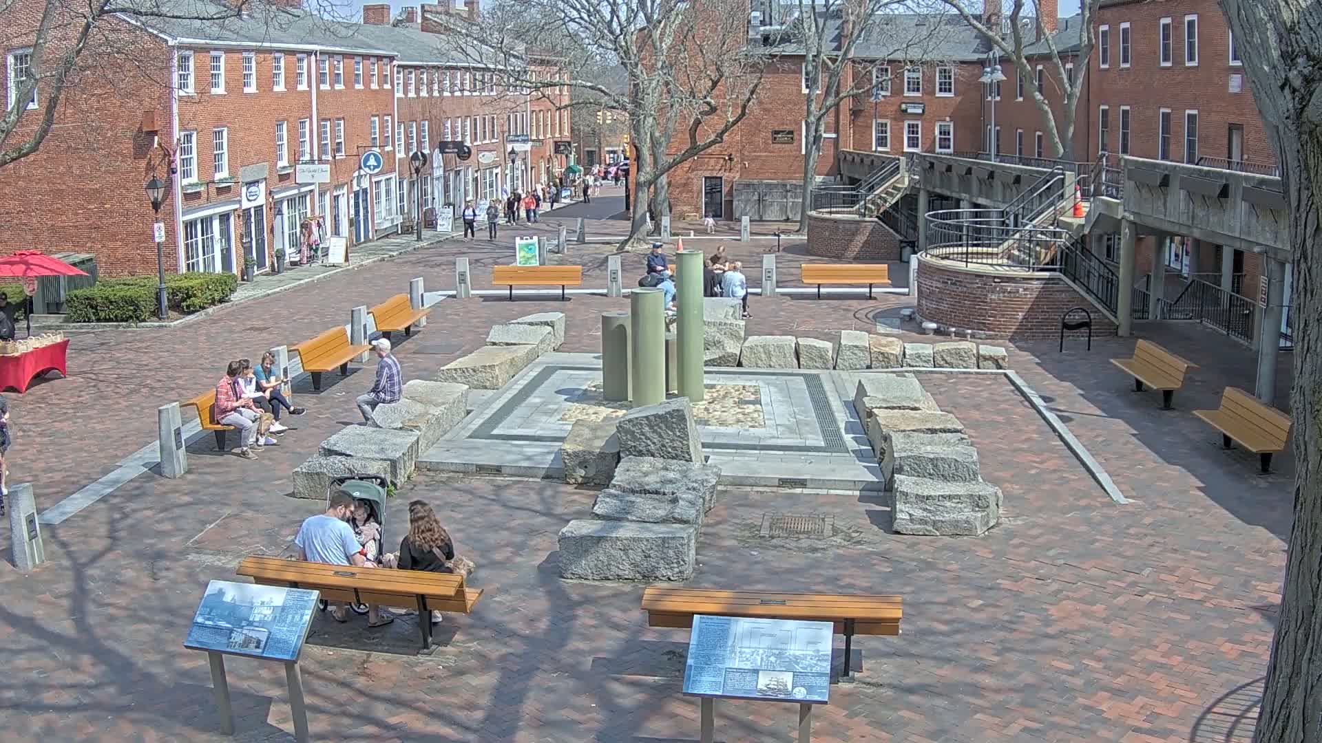 A brick plaza with a stone fountain is surrounded by brick buildings and several people are sitting on benches on a sunny day.