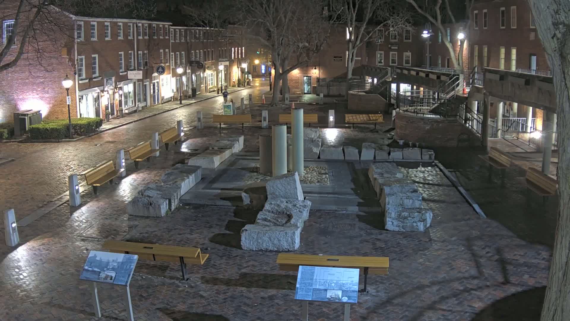 A brick plaza at night, featuring large stone ruins in the center, surrounded by benches and illuminated by streetlights.