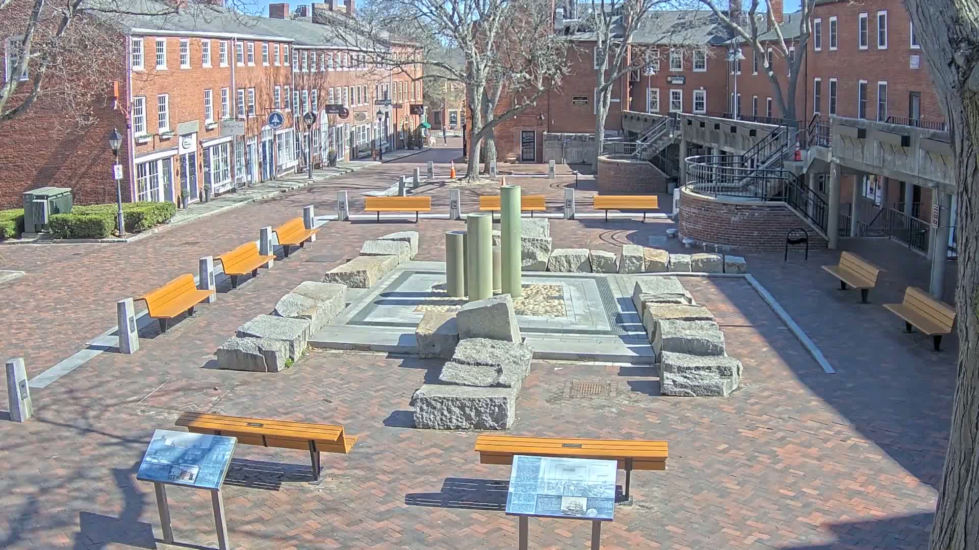 A brick plaza with several orange benches surrounds a stone fountain under a sunny sky.