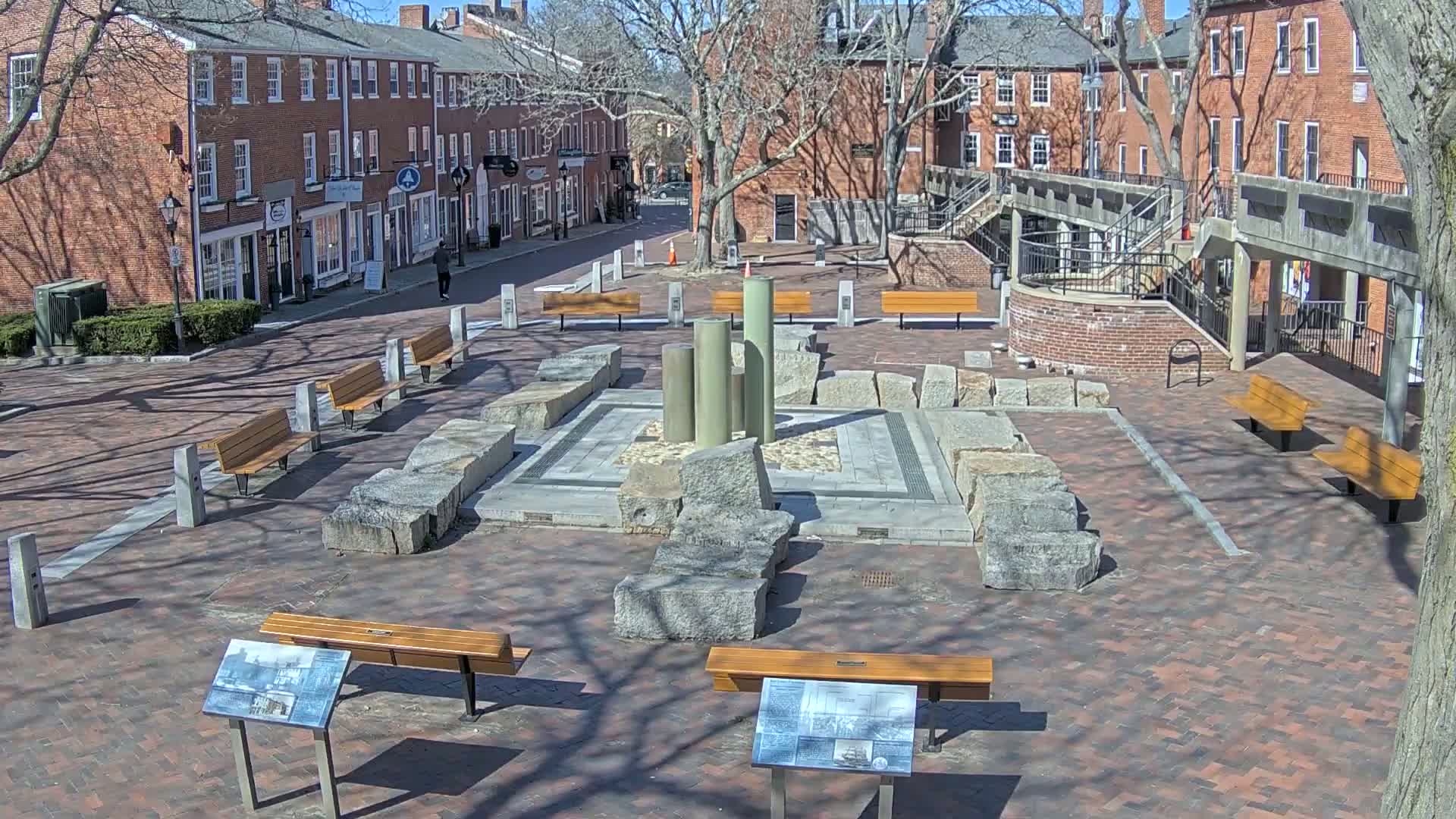 A brick plaza with several benches surrounds a stone and metal fountain, under a sunny sky.