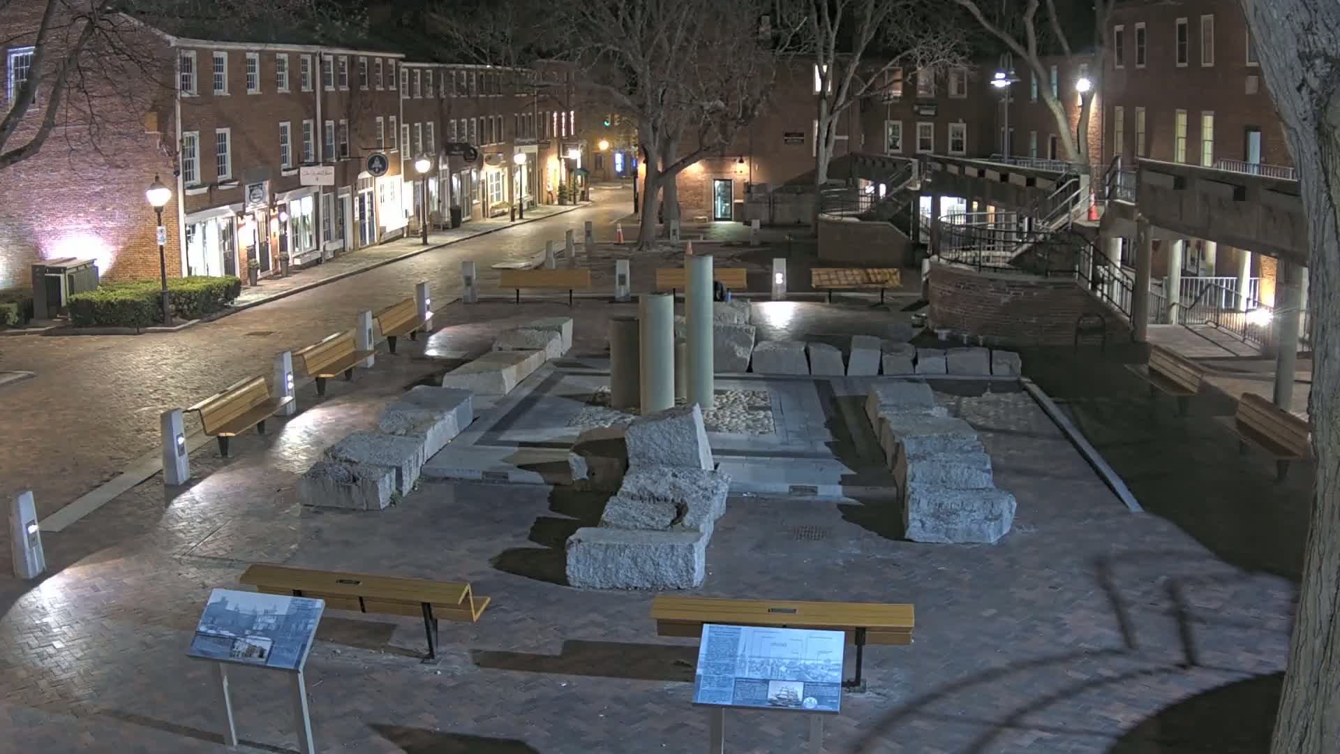 A nighttime view of a brick plaza with several benches and a central display of large stone ruins, illuminated by streetlights.