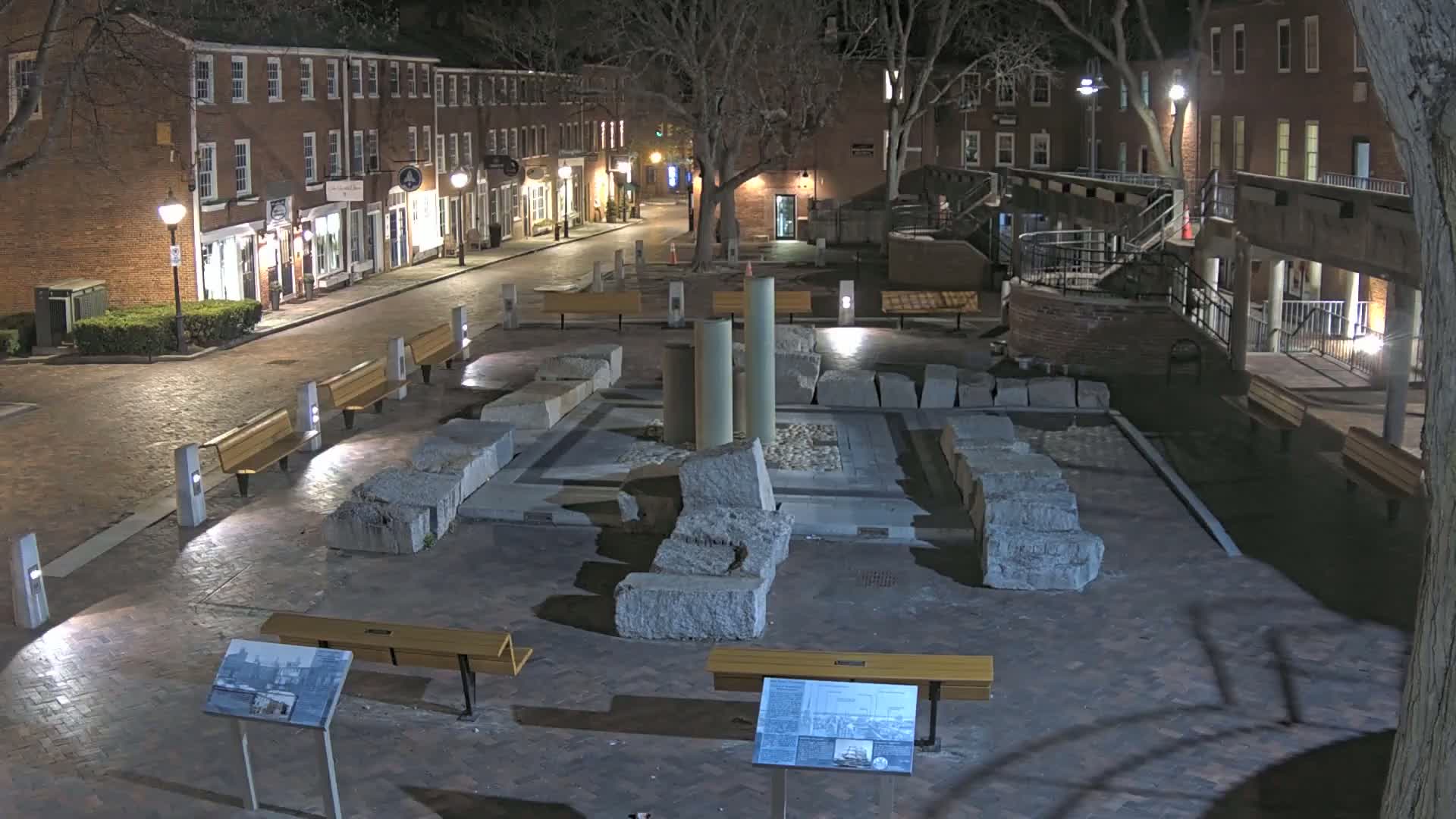 A nighttime view of a brick-lined plaza with stone ruins at its center, surrounded by brick buildings and benches under bare trees.