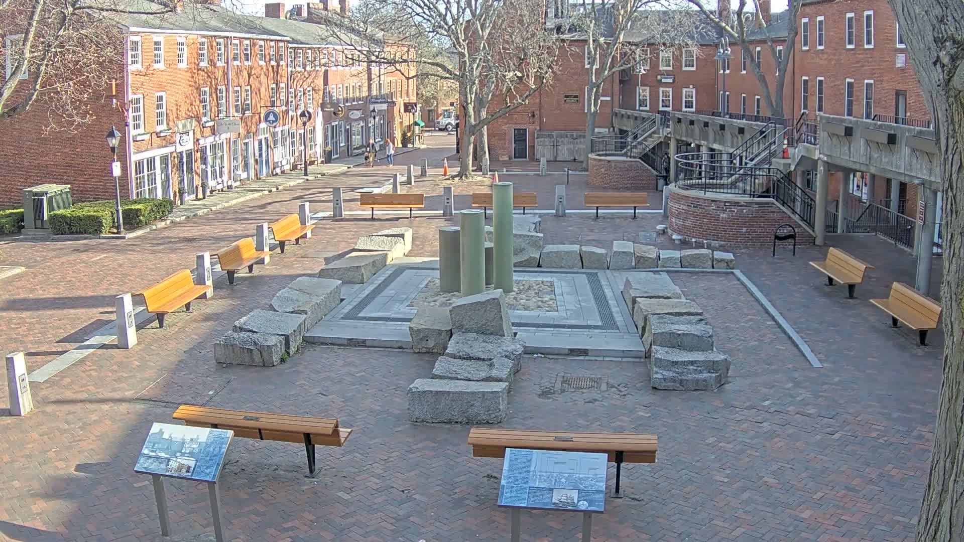 A brick plaza with several benches surrounds a central stone structure and fountain under a sunny sky.
