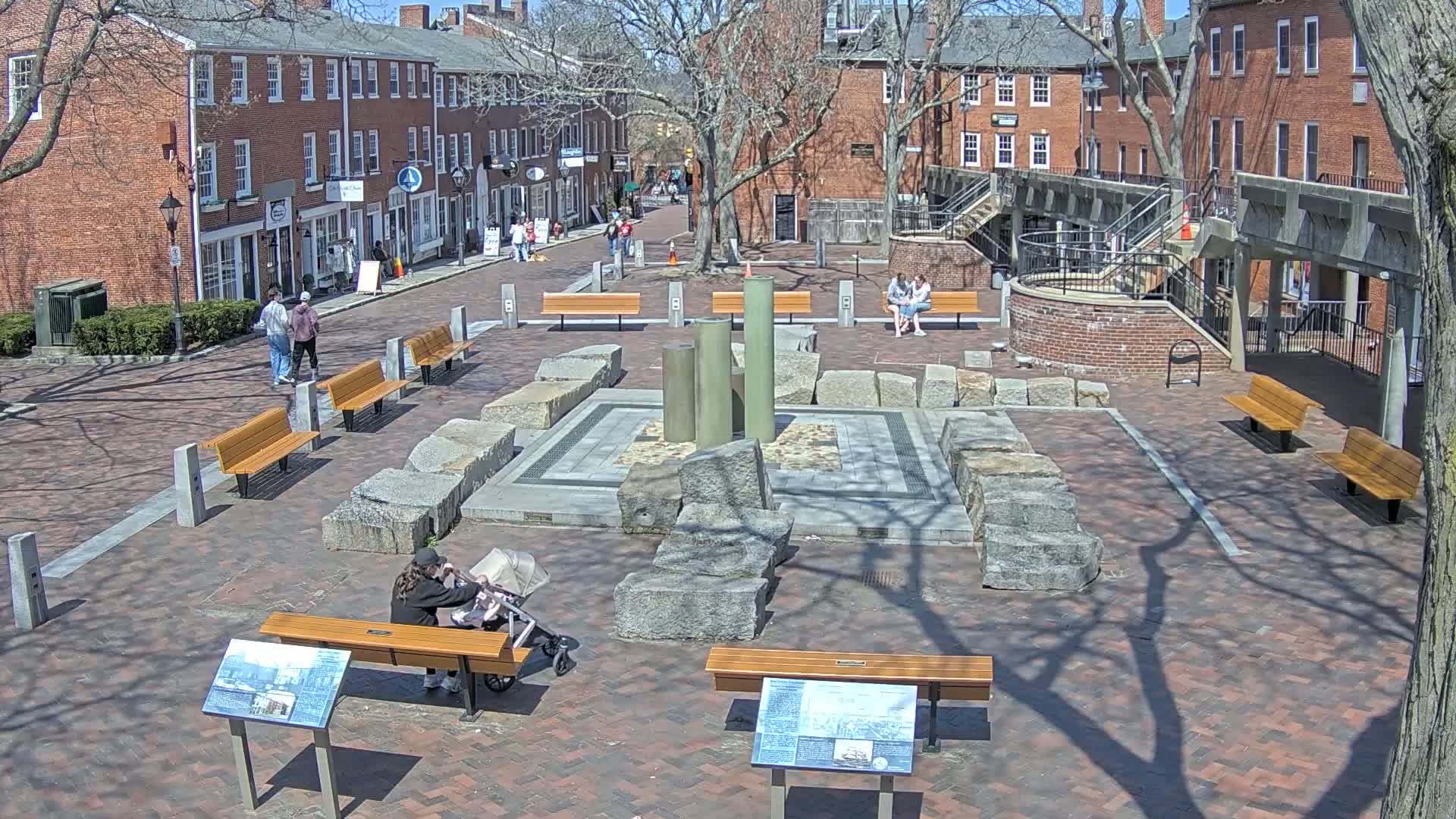 A sunny brick plaza with several benches, a stone structure in the center, and brick buildings surrounding it.