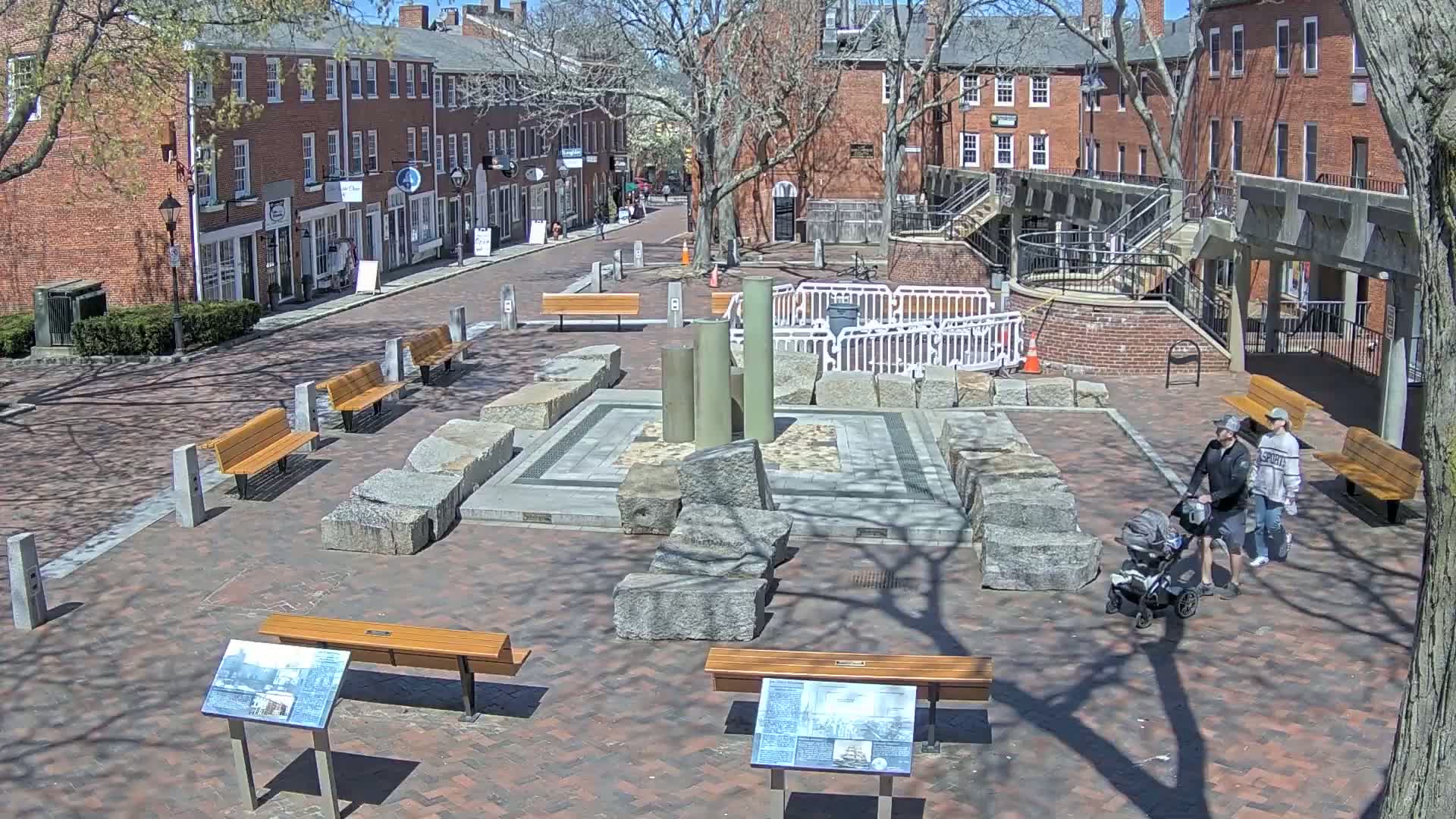 A sunny day in a brick-paved town square with several benches, a stone fountain structure, and two people pushing a stroller, near some historic-looking buildings.