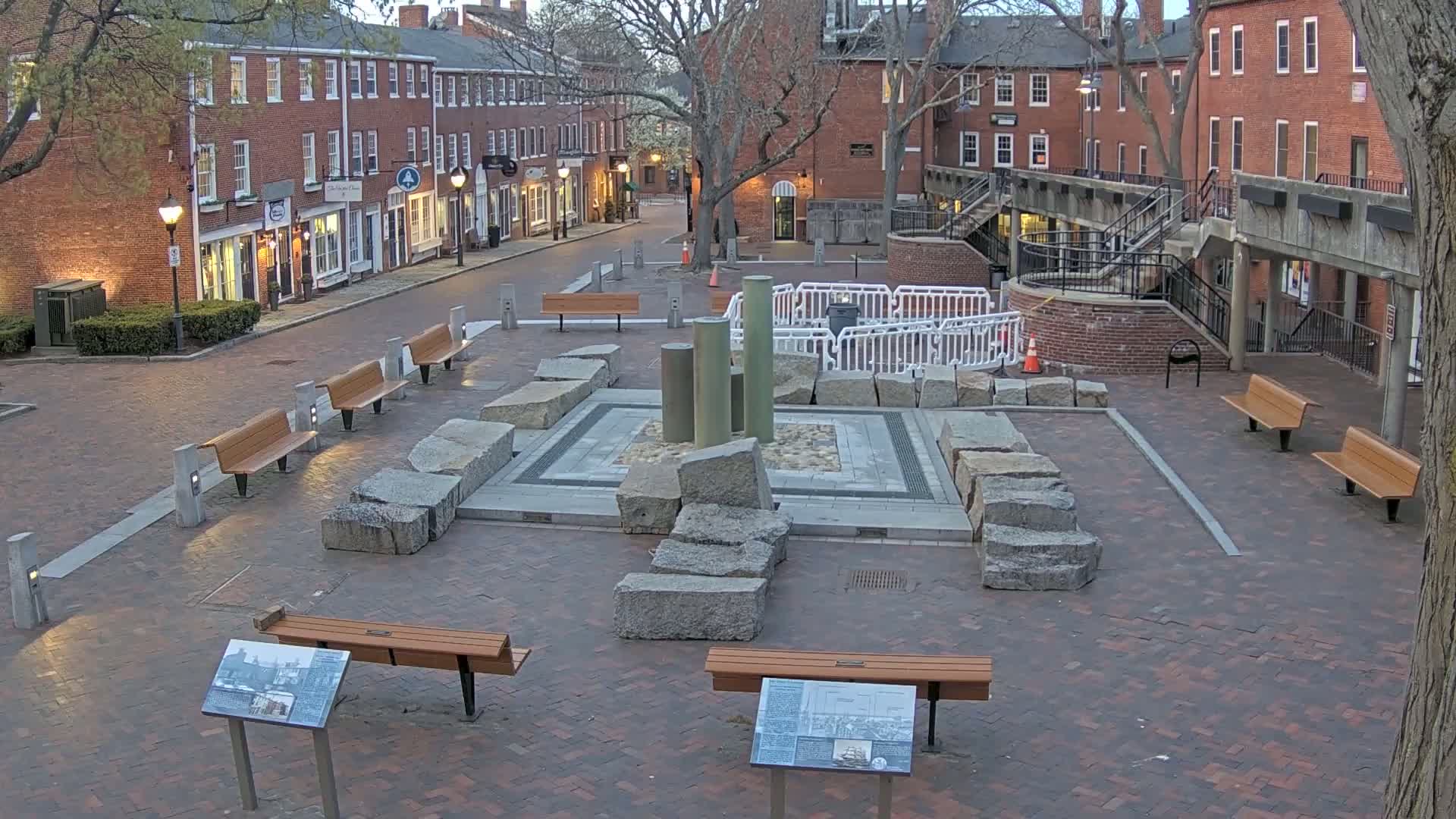 A brick plaza, under a partly cloudy sky, features a central stone structure with several benches and informational plaques surrounding it, and brick buildings lining a street beyond.