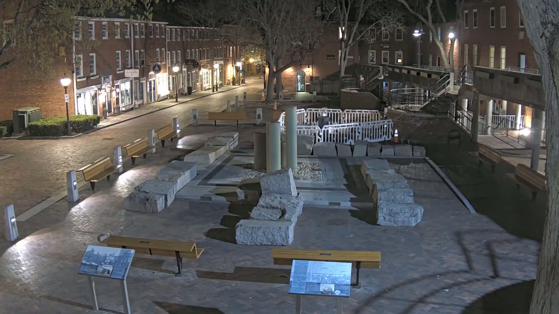 At night, a brick-paved town square contains remnants of a stone structure, surrounded by benches and lit by streetlights, with nearby brick buildings under leafless trees.
