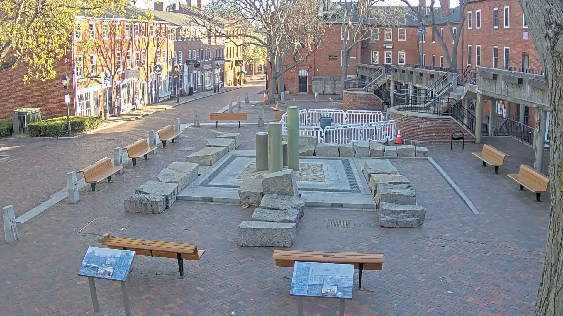 A brick town square with several benches, a stone fountain structure, and brick buildings lining the street on a sunny day.