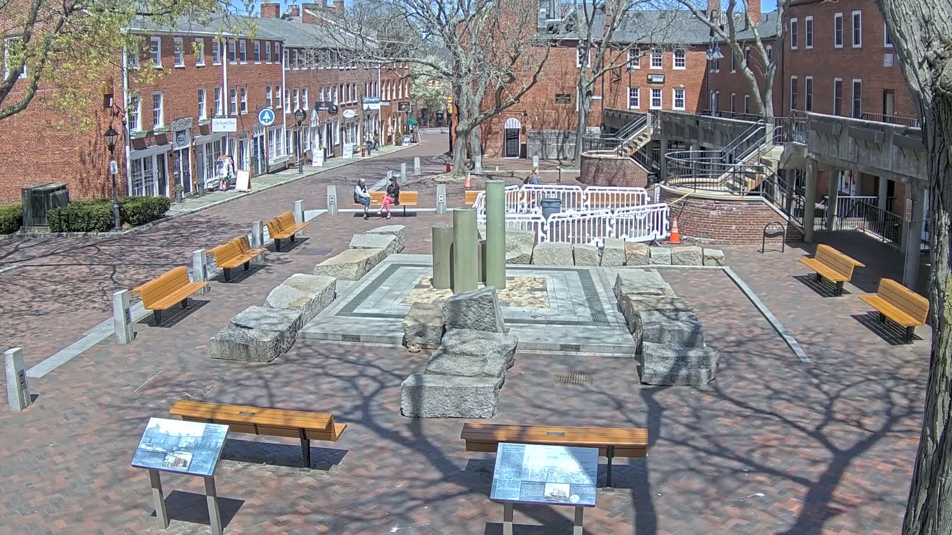 A brick town square on a sunny day features a stone fountain surrounded by benches and informational signs, with several brick buildings lining the square.